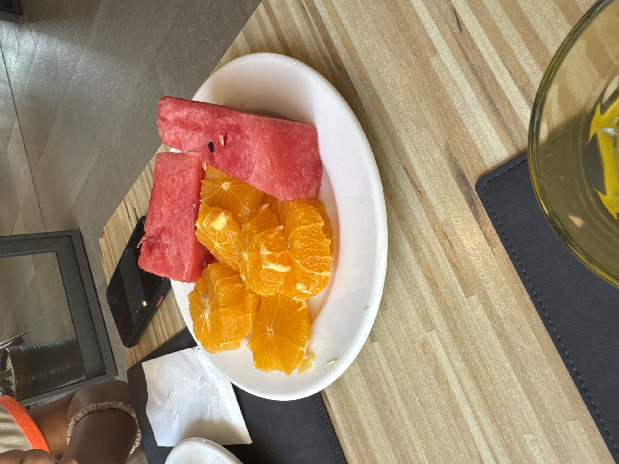 A plate of sliced oranges and watermelon sits on a light-colored wooden table. To the left of the plate, a white paper napkin is visible. Behind the plate, a black smartphone rests on a dark placemat. In the background, a blurred reflection shows a potted plant and part of a room, including what appears to be a mirror and a person's legs wearing a light-colored top. A glass with a yellow liquid is partially visible in the foreground. The setting is likely a cafe or restaurant in Aksu, Turkey, given the context provided.