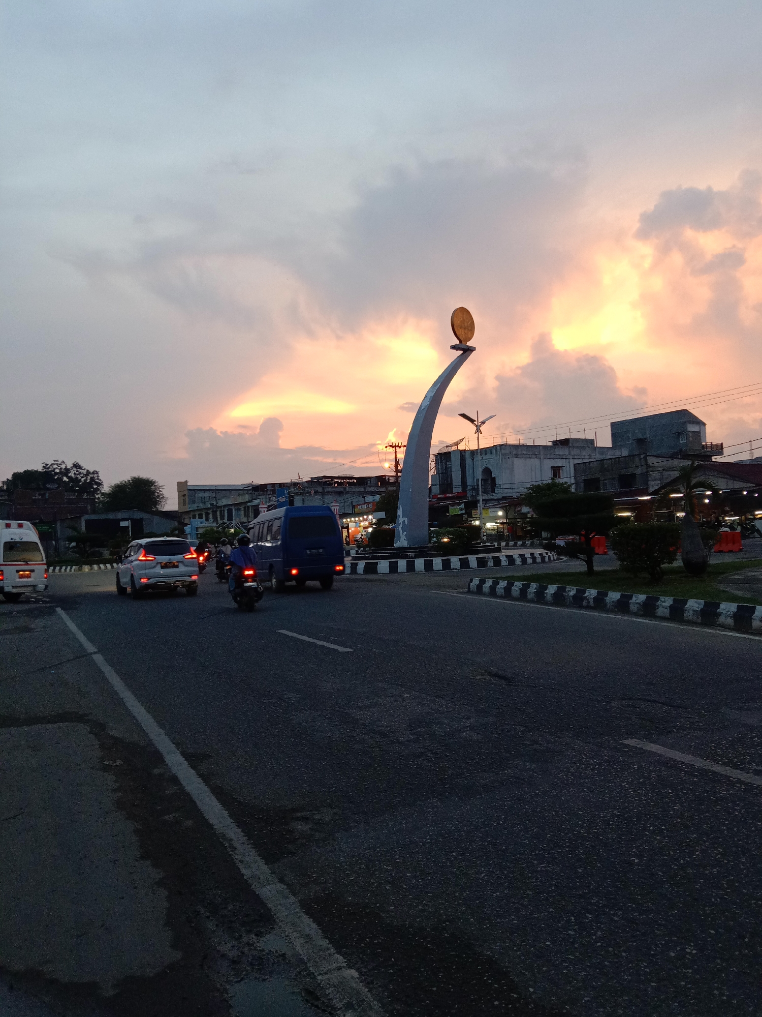 This image captures an urban roundabout at dusk in Lhokseumawe, Indonesia, characterized by a striking sunset sky. The scene is illuminated by the fading daylight, casting a warm glow over the clouds and creating silhouettes of the cityscape.
In the foreground, a dark asphalt road occupies the lower portion, with visible white lane markings. Traffic is active, featuring several vehicles: a white minivan on the far left, a silver sedan with illuminated taillights, a dark blue minivan further right, and multiple motorcycles with riders navigating between the cars.
The central feature is a prominent monument located within the roundabout. This monument is a tall, curved, grey structure ascending from the ground, topped with a circular, golden disc. The roundabout itself is landscaped with small shrubs and trees, bordered by distinct black and white striped curbs. Surrounding the roundabout and lining the streets are various buildings, some appearing to be commercial or residential, with dimly visible lights from businesses. Overhead, power lines crisscross the sky.
The upper half of the image is dominated by the sky, displaying a blend of soft blue, grey, and vibrant orange-yellow clouds, indicative of sunset. The light is diffused, creating a serene yet busy atmosphere as the city transitions into night. No discernible text is clearly visible on any signs or structures.