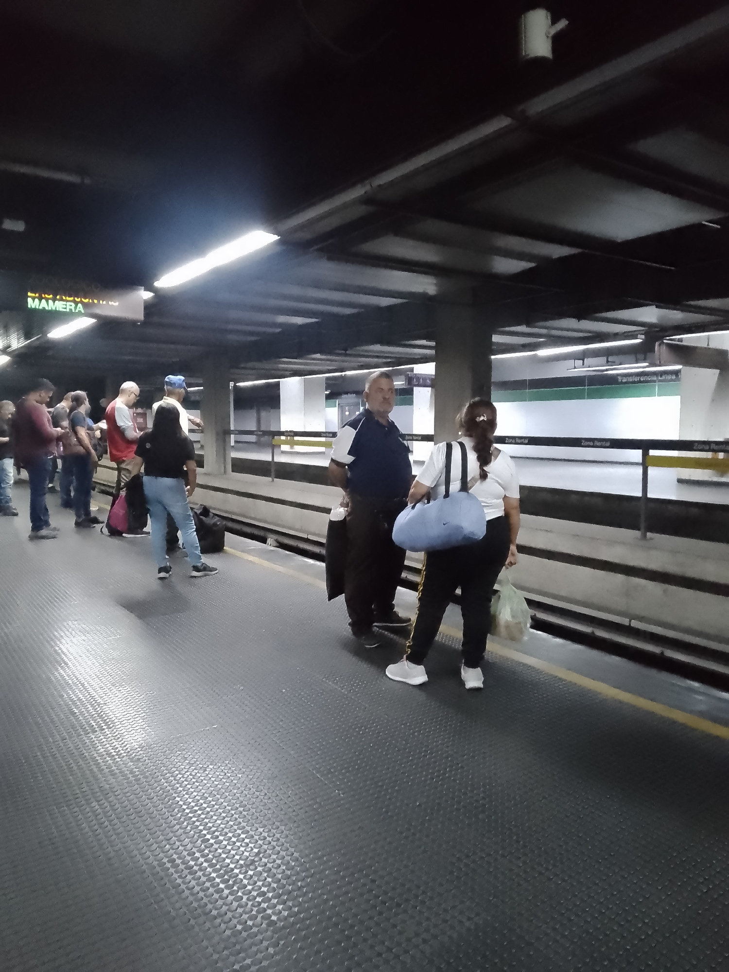The image depicts a subway station platform, likely in Caracas, Venezuela, evidenced by the sign “Las Adjuntas Mamera” visible in the upper left corner. A group of people stands on the platform, waiting. A man in a dark blue shirt and dark pants stands with a woman in a white shirt and dark pants who carries a light blue duffel bag and a plastic bag. Signage behind them indicates "Transferencia Linea 1". The platform has a textured, dark gray surface and a yellow safety line.