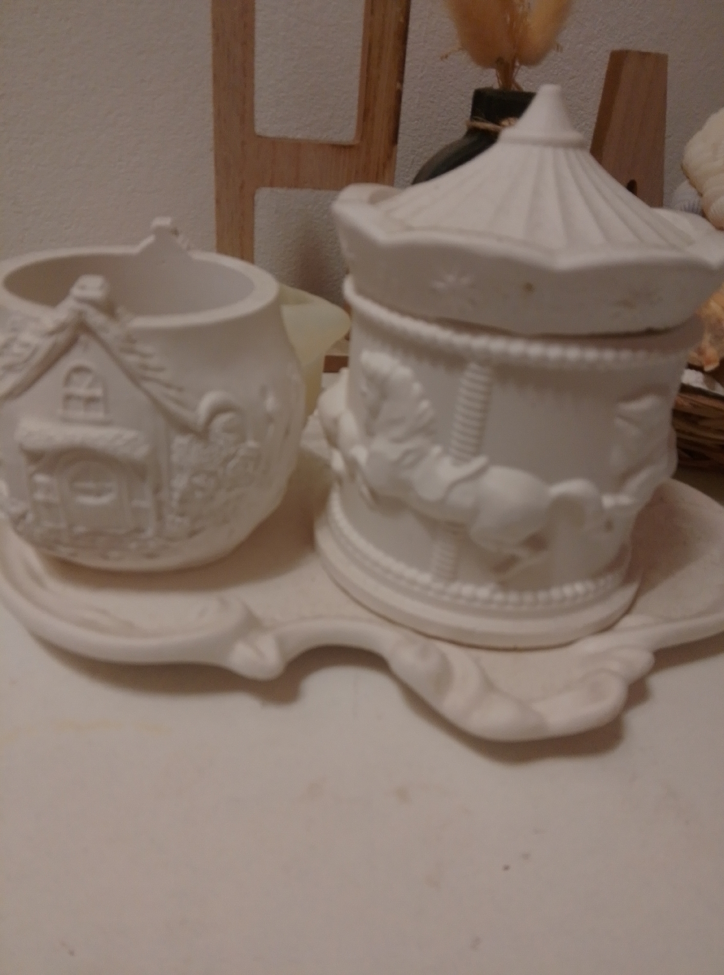 This is a photograph of two unpainted ceramic figurines on a white surface. The figurine on the left is a small, round pot shaped like a cottage with a pitched roof, a window, and a door. The figurine on the right is a carousel with horses around its base and a conical, tent-like lid. Both figurines are placed on a white, decorative tray. The background includes a wooden structure and some dried plant material. The image does not contain information about the city of Wedel, Germany.