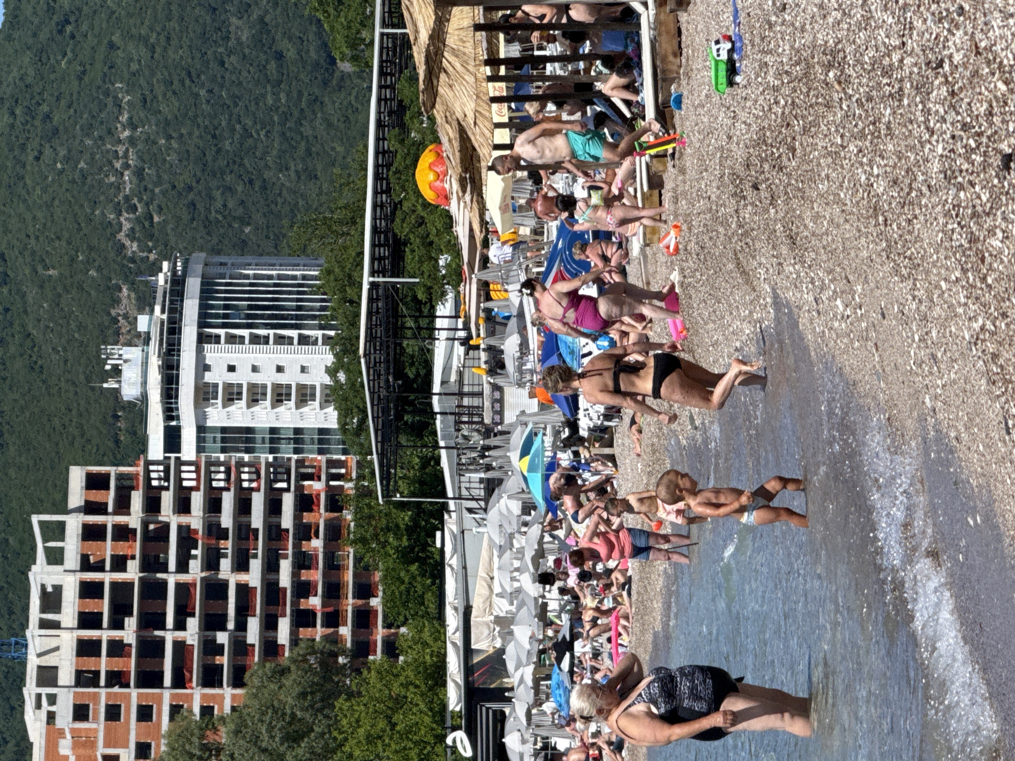 This image depicts a bustling pebble beach scene in Boreti, Montenegro, under bright daylight.
In the background, a dense green forested mountain dominates the upper portion of the frame. Below the mountain, two prominent multi-story buildings are visible. To the left, a large, unfinished construction project shows a concrete skeleton with exposed brickwork and red safety netting within window openings. A crane is partially visible above its roofline. To the right, a modern, finished white high-rise building with numerous windows and balconies stands, featuring vertical architectural elements and rooftop antennae.
The mid-ground features extensive beach infrastructure. A large area behind the immediate beach is covered with rows of white and grey beach umbrellas over what appears to be a paved or decked surface. Several open-air, dark-colored structures, possibly restaurant or bar facilities, are present. Closer to the water, straw-thatched beach umbrellas shade multiple loungers on the pebble beach. A large, orange and yellow inflatable structure is visible further down the beach towards the right. A "Coca-Cola" logo is discernable on one of the straw umbrellas.
The foreground is dominated by the pebble beach and numerous individuals. The beach comprises small, light-colored pebbles, with a wet strip at the waterline and dry pebbles further up. The water is clear and light blue, showing small breaking waves.
Numerous people of varying ages and body types are present, engaged in typical beach activities. In the immediate left foreground, an older woman with light hair, wearing a black and white patterned one-piece swimsuit, stands in the shallow water. In the center foreground, a young child wearing a white swim diaper wades in the water, facing right. Slightly behind this child, a woman in a pink top and denim shorts stands, looking downwards. To the far right, a woman in a black two-piece swimsuit walks away from the camera, partially in the water. Other individuals are seen sunbathing on loungers or towels, standing, walking, and socializing across the beach. A man in light blue swim trunks walks towards the water on the right mid-ground. Several children are playing. Various beach toys, including a green and white toy truck, orange buckets, and a blue shovel, are scattered on the pebbles.