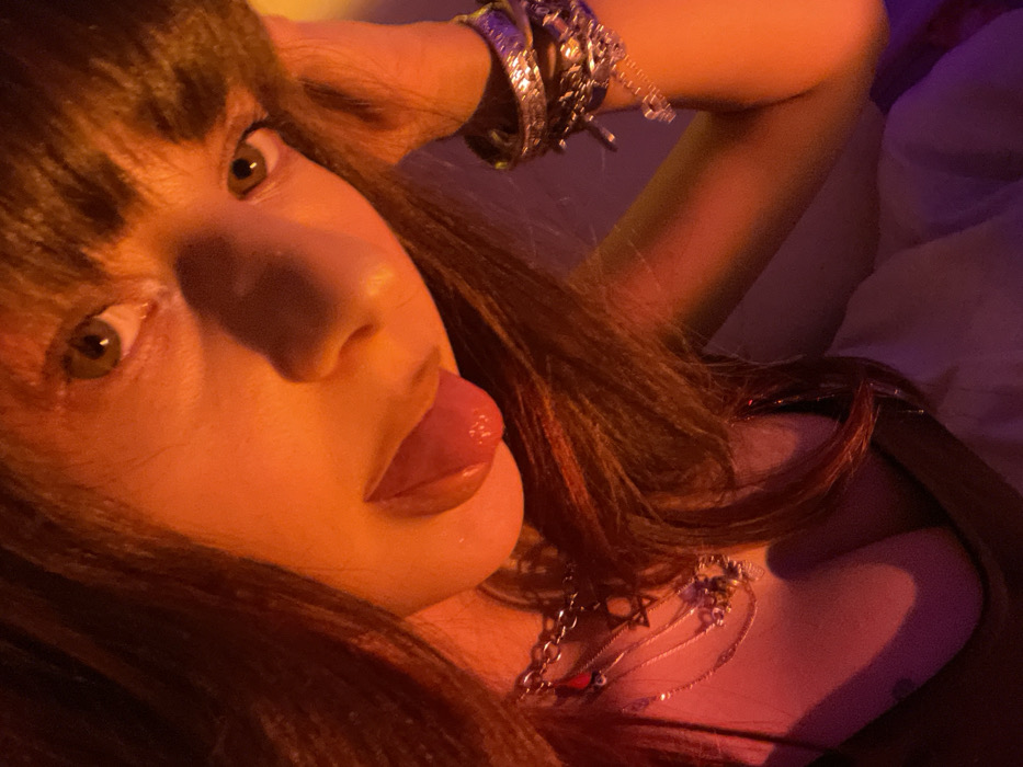 A close-up shot features a person with dark brown hair and bangs, lying down. The face, lit with an orange glow, is angled towards the camera, showcasing hazel eyes and a partially extended tongue. Multiple silver bracelets adorn their wrist, while a layered silver necklace rests on their chest. A hint of white fabric suggests a bed or pillow in the background. The overall aesthetic is intimate and suggestive.