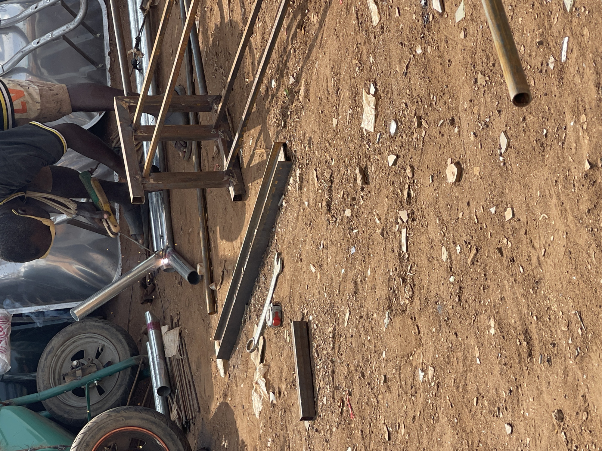 An outdoor scene in what appears to be a workshop or construction area in Abuja, Nigeria shows a man welding metal pieces together. The individual, wearing a welding mask, gloves and casual clothing is focused on joining metal pipes. The scene contains various metal tubes, rods, a ladder frame, a wrench, measuring tape, a car wheel, and various pieces of scrap scattered on a dirt surface. Welding sparks are visible at the point where the metals are being joined. The lighting suggests a sunny day.