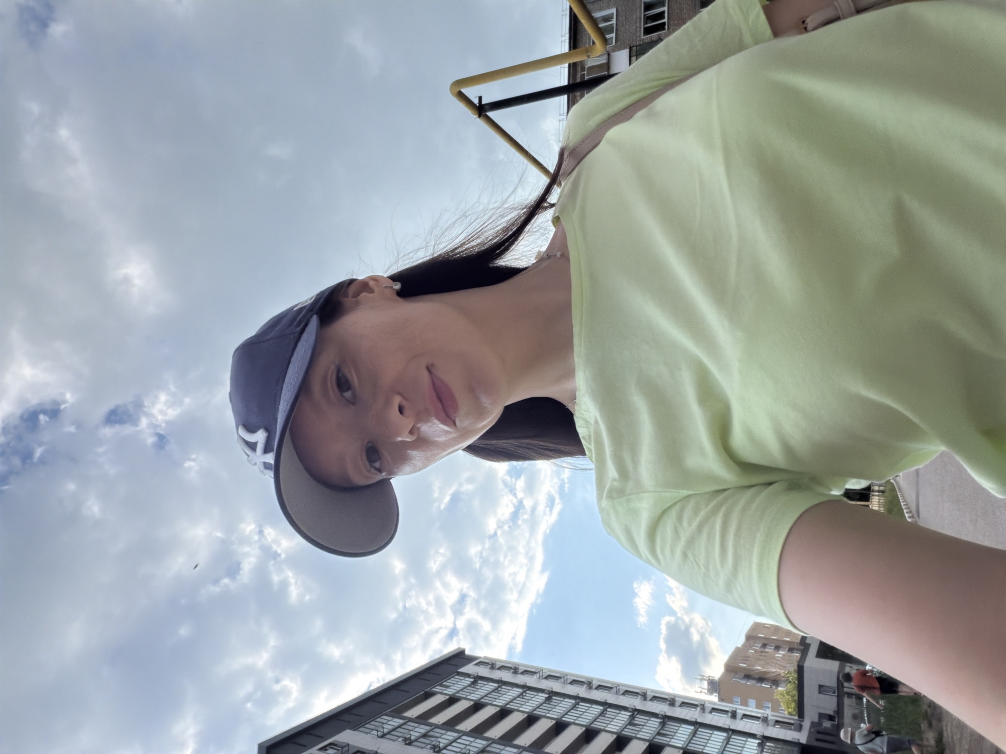 In the city of Tujmazy, Russia, a woman is captured in a selfie outdoors. She is wearing a light green t-shirt and a dark blue baseball cap. The background reveals a cloudy sky, a tall residential building on the left, and a playground structure on the right. A few other figures, appearing to be adults, are visible in the distance on a paved walkway. The woman's expression is neutral.