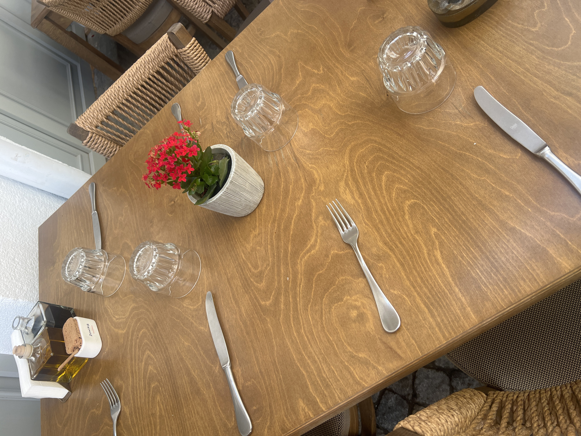 The image displays an overhead view of a set dining table, likely at an outdoor cafe or restaurant in Plovdiv, Bulgaria. The table is made of light brown wood, showcasing its natural grain.
The table is set for at least two diners. Each setting includes a silver-colored fork and knife, along with two clear, inverted glass tumblers. Centrally placed is a small grey, textured pot containing a vibrant plant with numerous small red flowers, characteristic of a Kalanchoe. To the lower left, a condiment holder is visible, featuring two glass bottles (one appearing to contain olive oil) and a small white ceramic dish with a porous brown block, possibly for dipping. The word "DUNAS" is clearly visible on the side of this white dish.
In the background and lower right, parts of the chairs are visible, characterized by their woven natural fiber (rope) backs and seats, suggesting a rustic or outdoor dining aesthetic. A light-colored wall or building facade is partially seen in the upper left background. The overall lighting is bright and even, indicating it is daytime. No people are present, but the arrangement implies the table is ready for guests or has recently been used.
