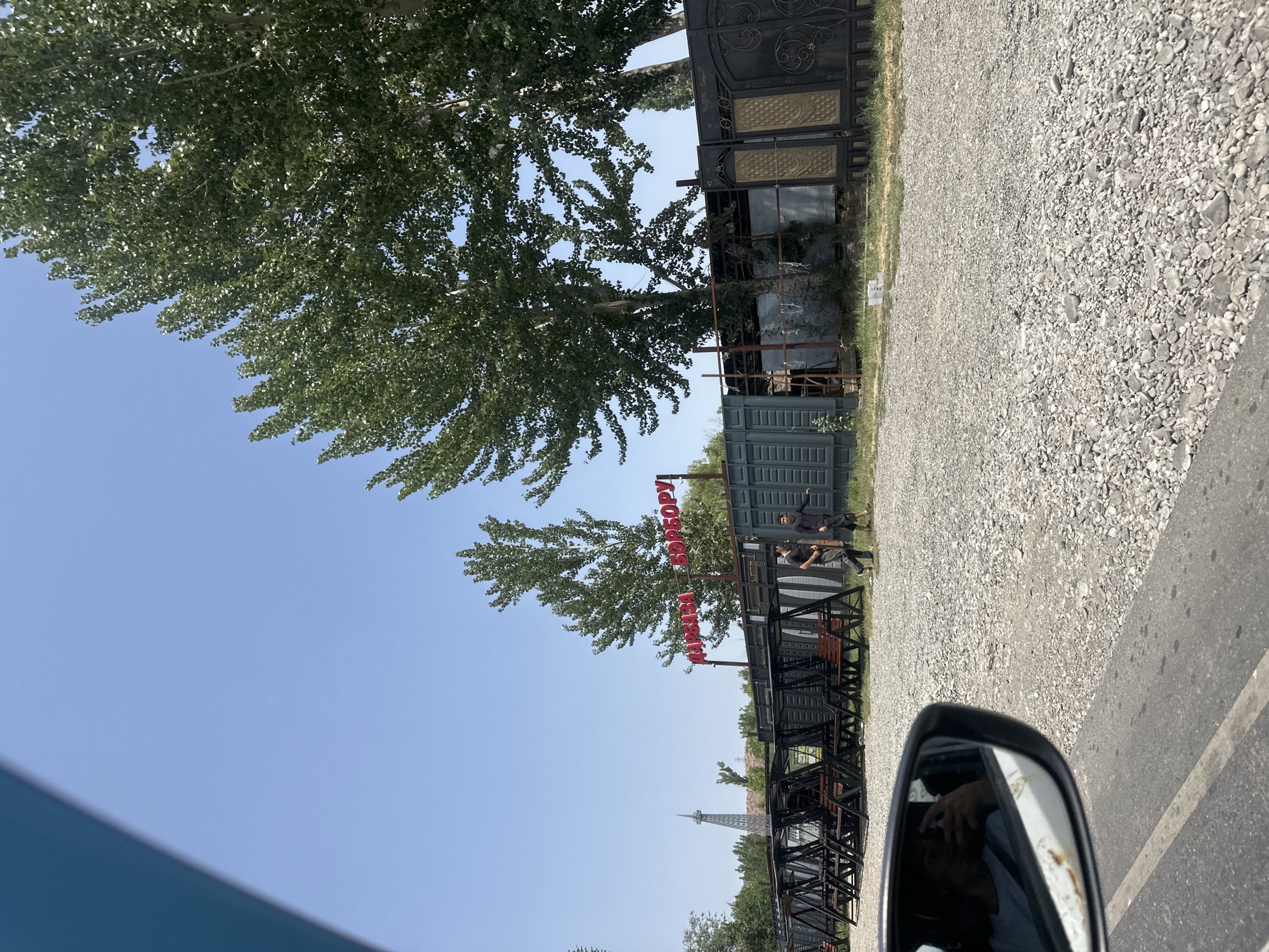 A view from a vehicle shows a street in Suzak, Kyrgyzstan, with a gravel lot, several empty swings, and a sign with red lettering that appears to read "ДАРВАЗА БОРБОРУ". Two men, one in a black shirt and the other in a black tracksuit, walk past the swings. Tall trees line the background, and a distant tower resembling the Eiffel Tower is visible on the left. The sky is clear and blue. The driver's reflection is visible in the car's side mirror.