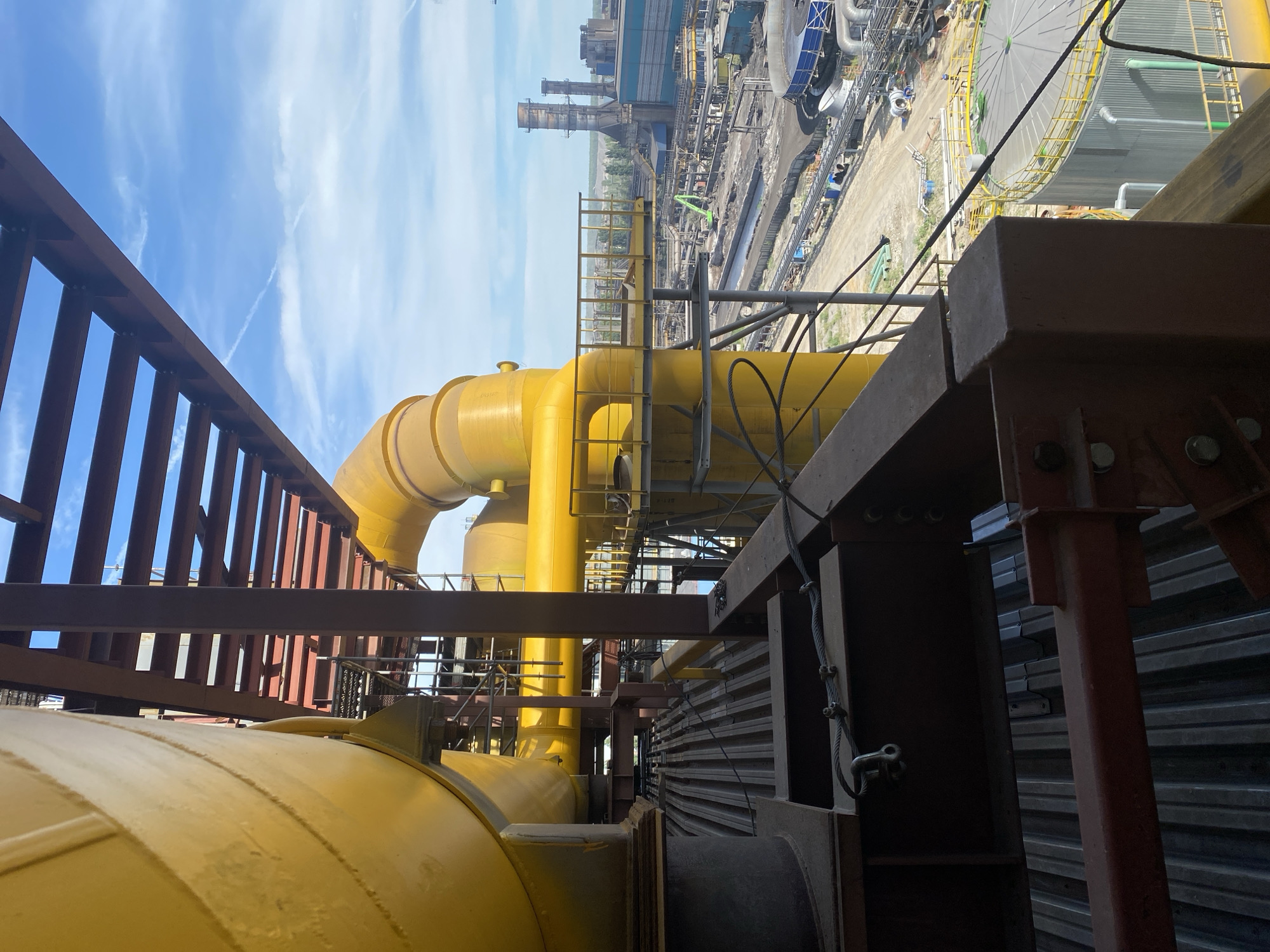The image depicts an industrial facility, likely a steel plant, in Lipetsk, Russia. In the foreground, large yellow pipes, part of an extensive industrial infrastructure, dominate the frame. These pipes are supported by a complex network of dark red structural steel beams and beams. The ground level consists of corrugated metal sheeting.
In the midground and background, the vast expanse of the industrial complex is visible under a clear blue sky with scattered white clouds and a faint contrail. The site features numerous industrial buildings, smokestacks, large tanks, and a network of smaller pipes and conduits. A bright green excavator is visible in the middle distance. Several figures, appearing small due to the distance, are scattered throughout the industrial grounds, engaged in various activities. The overall impression is one of large-scale industrial operations.