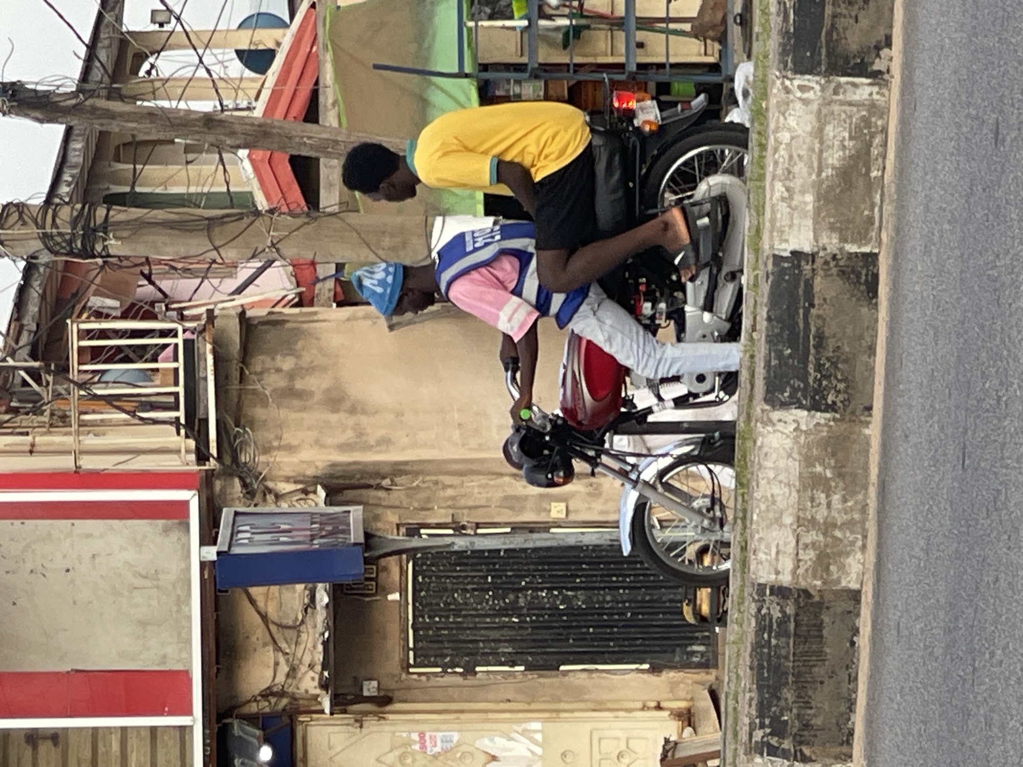 The image shows two individuals on a motorcycle, with the motorcycle parked on a street. The person in front is wearing a pink shirt, a blue vest with white lettering, and a blue cap with white markings. They appear to be in a seated position, potentially driving or about to drive the motorcycle. The person behind them is wearing a yellow t-shirt and black shorts, and is also seated on the motorcycle.
The background is somewhat cluttered, suggesting an urban or semi-urban environment. There are buildings with exposed wiring and what appears to be a metal grate or shutter on one of the structures. A portion of a road is visible on the right side of the image, with a pedestrian sidewalk or curb featuring a black and white striped pattern. The lighting suggests it is daytime, possibly overcast. There is no clear indication of the specific location or the weather conditions beyond it being light enough to see. The overall scene appears to be a candid shot of daily life on a street.