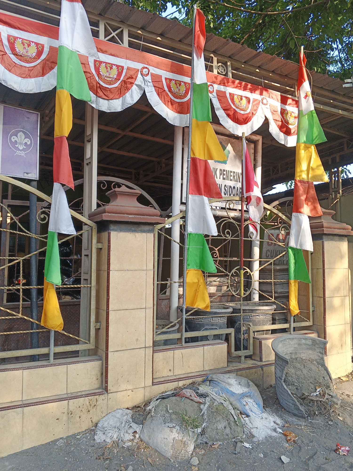 The image depicts the exterior of a building in Krian, Indonesia, likely a public facility. A red and white bunting, featuring the Indonesian coat of arms, is draped along the roofline. Numerous small flags in red, white, green, and yellow are hanging from poles and fences, suggesting a national celebration or event. A sign with Indonesian text is visible behind the flags. The foreground shows a sidewalk or paved area with debris, including discarded materials and a broken planter. The overall scene suggests a slightly neglected but festive outdoor space.