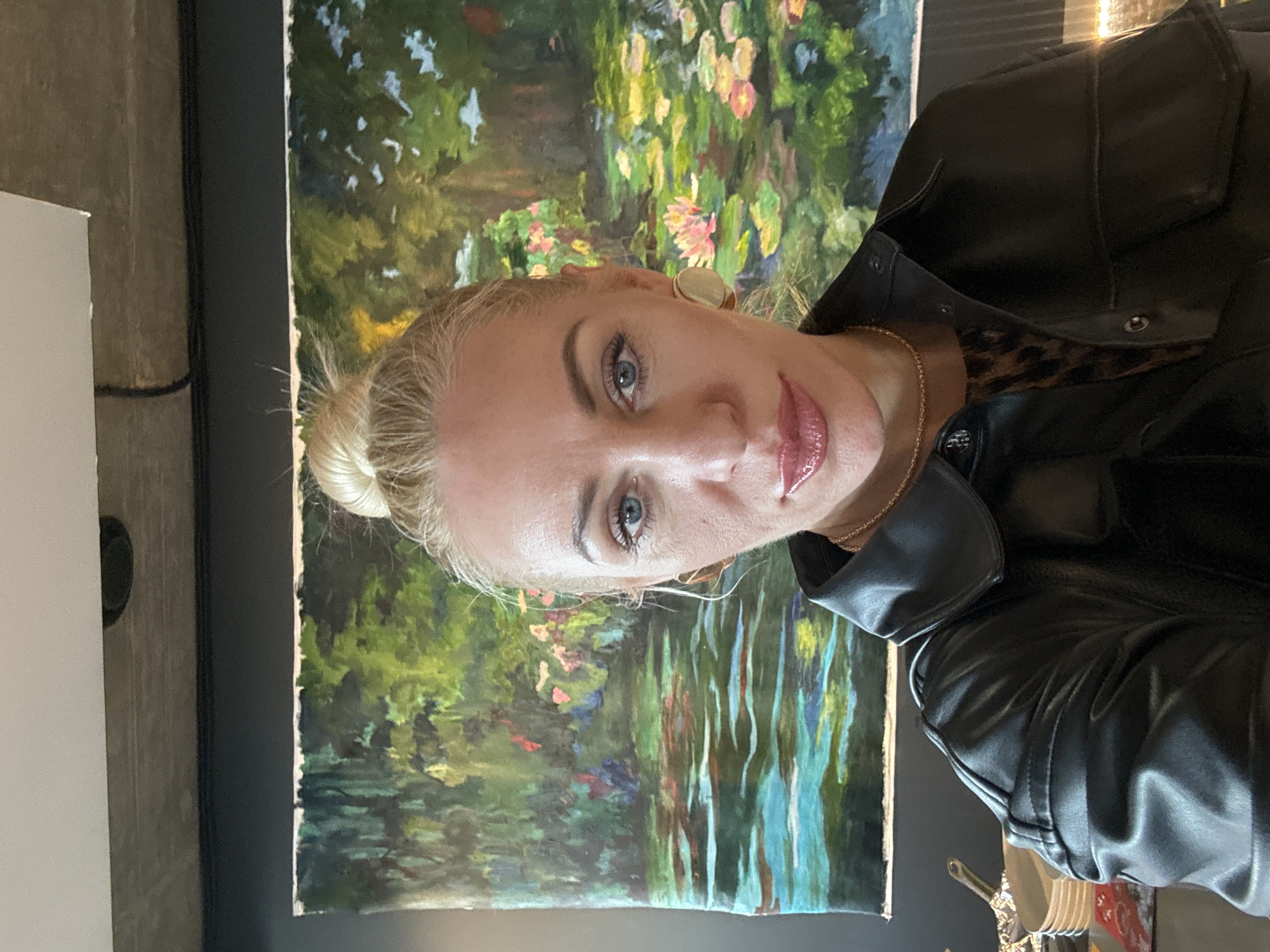 The image presents a close-up indoor selfie of an adult female. She has blonde hair pulled into a high bun, blue eyes, and is wearing visible makeup including eyeliner, mascara, and glossy pink lip color. She wears a black collared faux leather button-down shirt-jacket, with a leopard print top visible underneath. Her accessories include a thin gold chain necklace and gold circular stud earrings. She maintains a direct gaze at the camera with a neutral expression.
Behind her, a large, horizontal painting is displayed on a dark gray wall. The unframed artwork depicts an impressionistic water scene, resembling a pond with green foliage, reflective turquoise water, and scattered pink water lilies. Above the dark wall, a light-colored concrete ceiling is visible, featuring dark linear elements and a circular recessed fixture. In the lower left background, partially obscured, are a stack of white plates and metallic kitchen utensils. A partial light source is visible in the lower right background. No visual information within the image provides evidence to identify the location as Saint Petersburg, Russia.