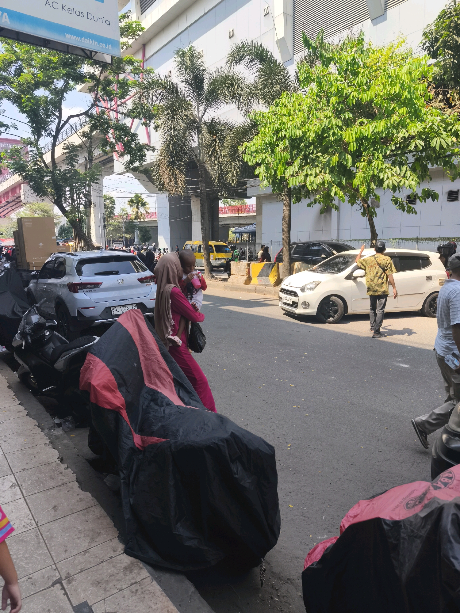 The image depicts a street scene in Kelurahan 16 Ilir, Kec. Ilir Timur I, Indonesia, on a sunny day. A light-colored Honda WR-V SUV is parked on the side of the road. A woman wearing a pink dress and a beige headscarf is standing next to the SUV, holding a young child. Several motorcycles are parked on the sidewalk and covered with tarps. In the background, a white sedan is visible, and a man in a camouflage shirt is standing near it, with his back to the camera, raising his hand. Other individuals and vehicles are present further down the street, and trees and overhead structures suggest an urban environment. A billboard with text including "AC Kelas Dunia" and "www.daikin.co.id" is also visible.