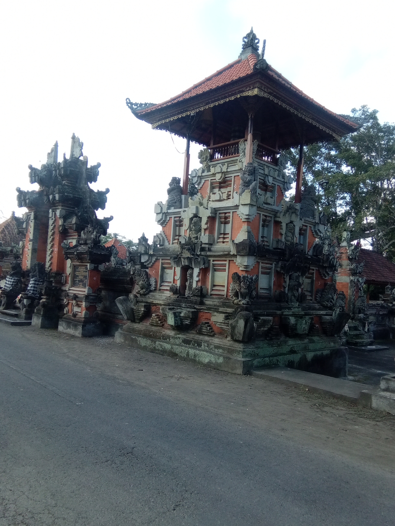 In an outdoor scene, an ornate Balinese temple gate stands adjacent to a paved road. The temple's architecture features intricate stone carvings, multiple levels, and a tiled roof. Statues of mythical creatures and figures adorn the structure. The road is empty, and a large tree with dense foliage is visible behind the temple. The sky is overcast.