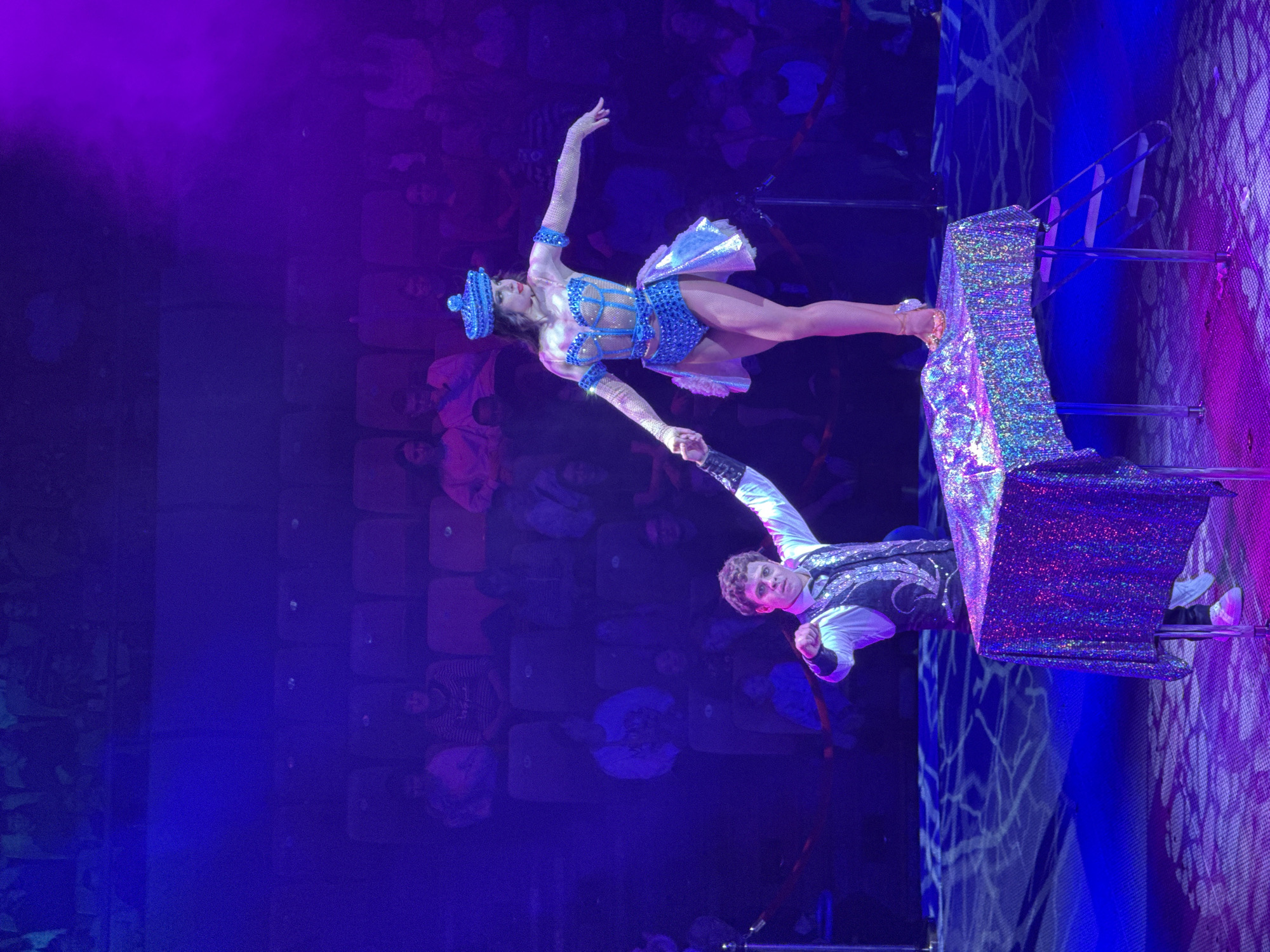 Two circus performers are captured mid-performance, lit by vivid blue and purple lights, against the backdrop of a seated audience. The male performer, wearing a white shirt and patterned vest, stands on a raised, sparkling platform, holding the hand of the female performer who is suspended in the air. The female performer wears a blue, sequined costume with a matching hat. The location is identified as Izhevsk, Russia.