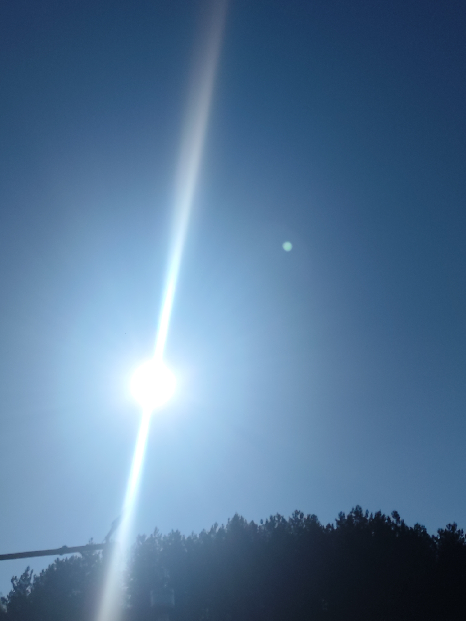 The image shows a bright sun in a blue sky, with a vertical streak of light extending from it. A line of trees is visible at the bottom of the image. A faint, small, green-tinted circle is in the upper-right quadrant of the sky. A pole is on the lower left side of the photo. The location is Unknown, Unknown.