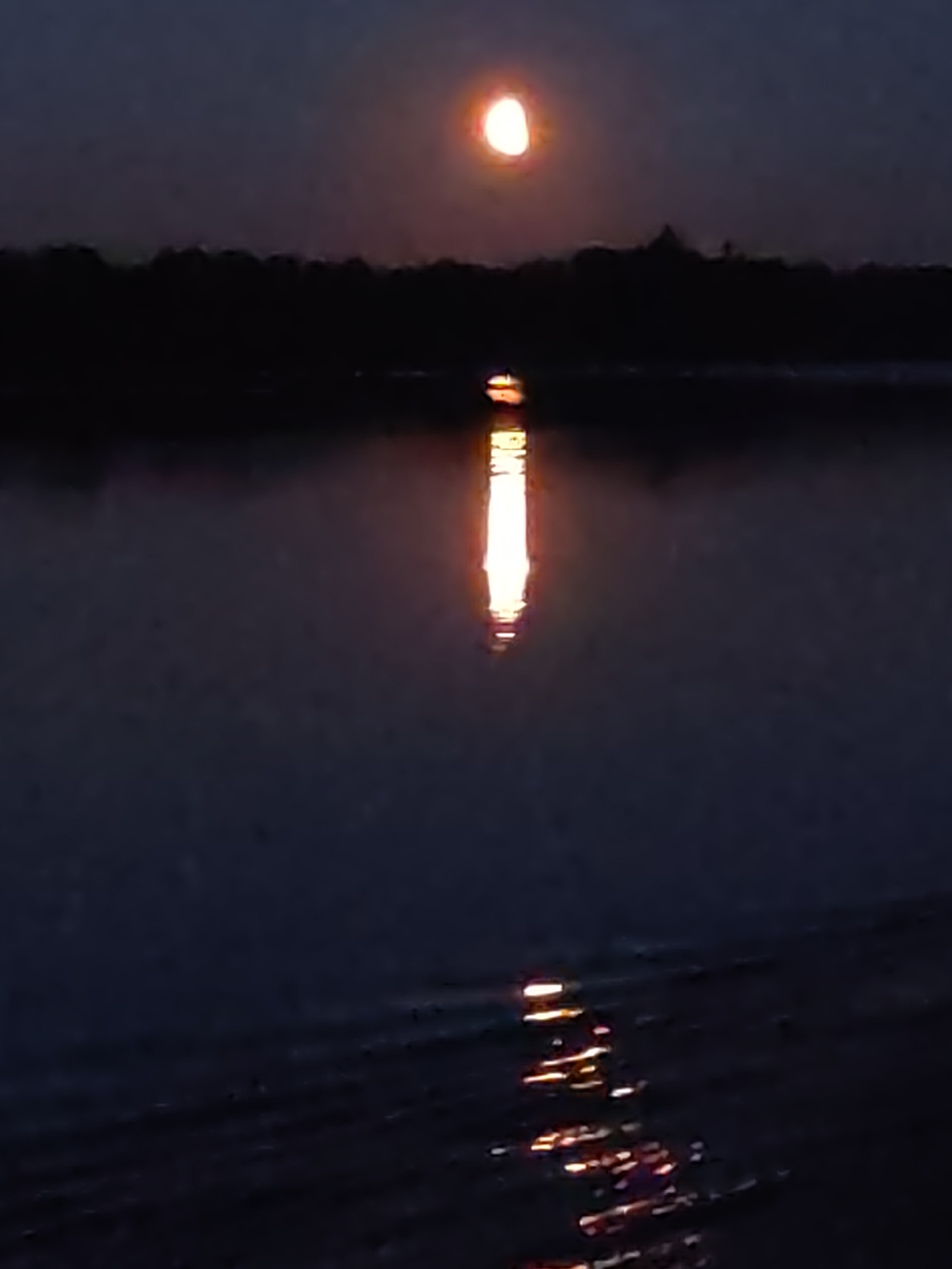 The image depicts a twilight or nighttime scene over a calm body of water in Yurino (Glebovskaya S/A), Russia. A single, luminous, orange-yellow celestial body, appearing slightly ovoid or crescent-shaped, is prominently positioned in the upper-left quadrant of the dark, dusky sky. Below this, a dark, silhouetted landmass, consistent with a dense tree line, extends horizontally across the middle ground, marking the distant shore.
The tranquil water surface directly reflects the celestial body, forming a distinct, elongated vertical column of bright orange-yellow light that originates near the horizon and extends downwards into the water. In the foreground, the water exhibits subtle ripples and small waves, which cause the reflected light to fragment into multiple shorter, scattered horizontal streaks of orange-yellow, varying in length and intensity.
The overall environment is natural and dark, with no discernible human subjects, artificial structures, or overt actions taking place. The scene is dominated by the interaction of natural light and water reflection against a dark landscape.
