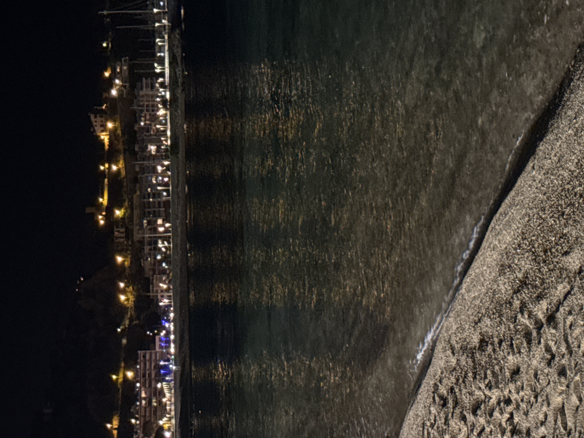 This is a nighttime photograph taken from a beach in Blanes, Spain, looking towards the coastline. The foreground features the sandy beach with gentle waves lapping at the shore. The water is dark and reflects the lights of the town in the background. The town itself is illuminated with numerous lights, indicating a populated area. Buildings are visible on the hillside, suggesting a coastal town built on an incline. Several boats with masts are docked in the water, likely in a marina. The overall impression is that of a lively coastal town at night, viewed from the peaceful setting of the beach.
