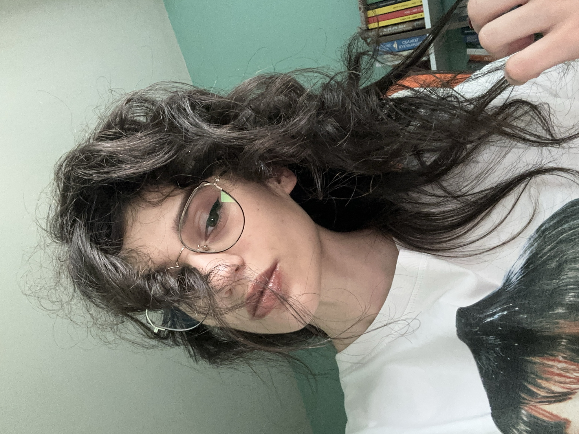 This is a portrait of a young person with long, dark, wavy hair and green eyes, wearing round, wire-rimmed glasses. The person is wearing a white t-shirt with a graphic print of a dog. Their lips are slightly parted and appear to have a glossy, pink-toned lip product. The person's hair is somewhat messy, with strands falling across their face. The background is a wall painted in two tones of green, with a bookshelf visible to the right, filled with books. The lighting suggests it is daytime, possibly indoors. The person's expression is neutral or slightly pensive. There is no visible text. The provided location context is Sofia, Bulgaria.