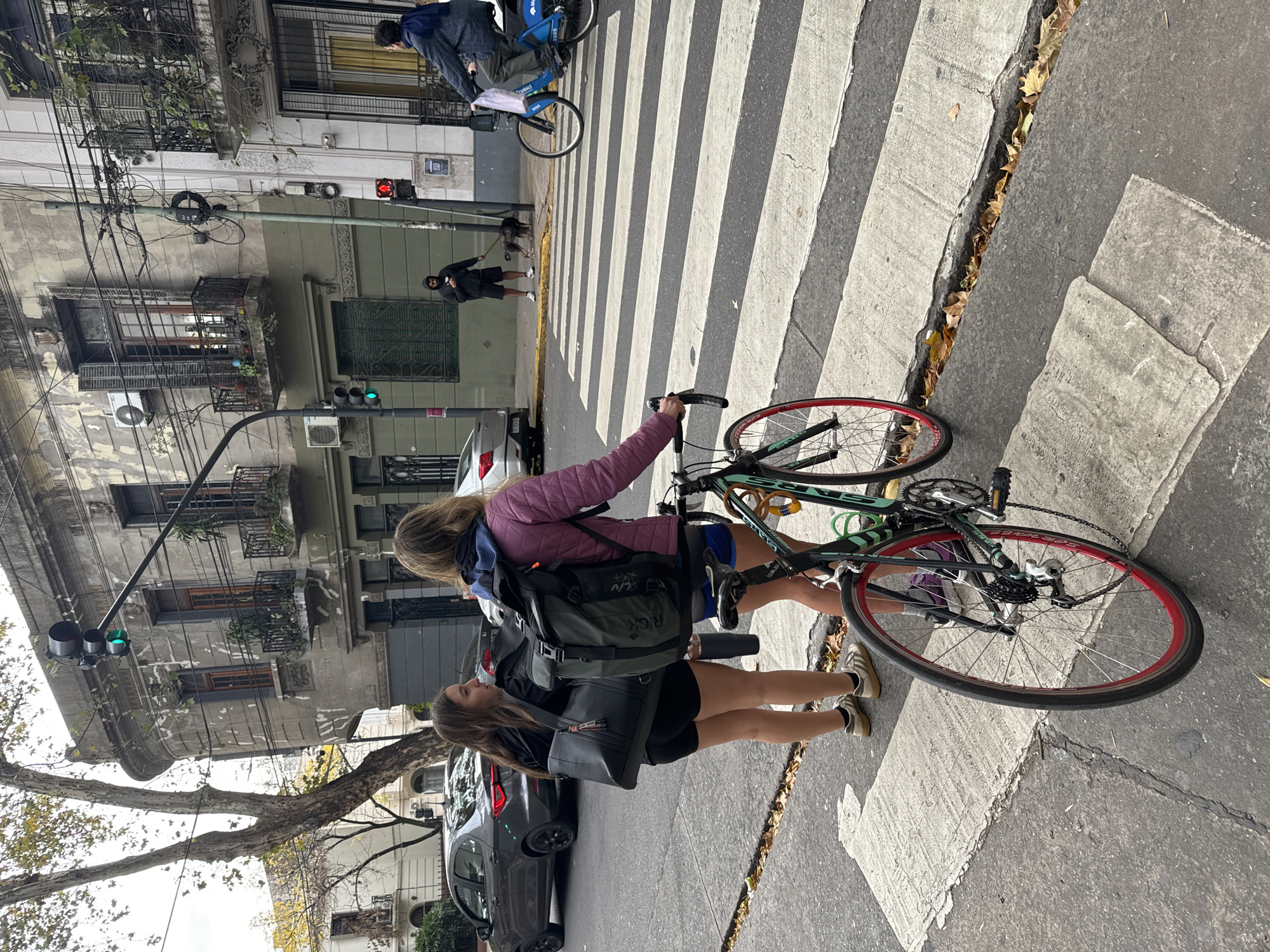 Two women with a bicycle are crossing a zebra crossing in Buenos Aires, Argentina. The building is a classic building in Buenos Aires, a tree, a car parked, and some other people crossing the street.