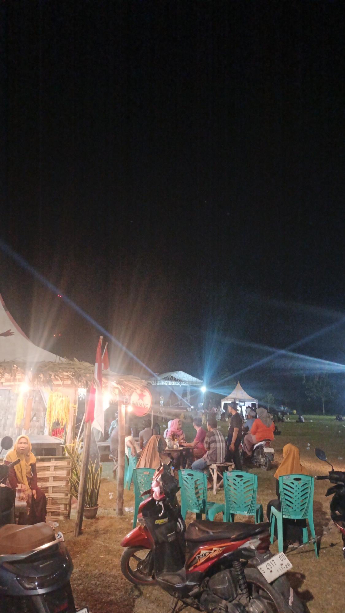 The image depicts an outdoor evening event at night, likely a festival or gathering, in Ketemasdungus, Indonesia. The scene is illuminated by artificial lights, including bright spotlights projecting beams into the dark sky and warm lighting from a stall or tent structure on the left. In the foreground, two motorcycles are parked on a grassy area. The one on the right is red and black, with a license plate reading "S 4917 OU". The one on the left is dark colored.
Further back, a group of people are seated at tables and chairs, some interacting with each other, while others are observing the event. The seating consists of bright teal plastic chairs and some wooden benches. A woman in a yellow hijab is seated on a wooden structure on the far left, facing towards the viewer. Other individuals are visible in the background, including some near a stage structure with lighting equipment. Flags, including what appear to be Indonesian flags, are displayed on poles near the stall. A circular sign with the word "BINTANG" is visible on the stall. Tents are set up in the middle ground, suggesting areas for vendors or seating. The overall atmosphere appears lively and social, with a clear distinction between the brightly lit event area and the dark night sky above.