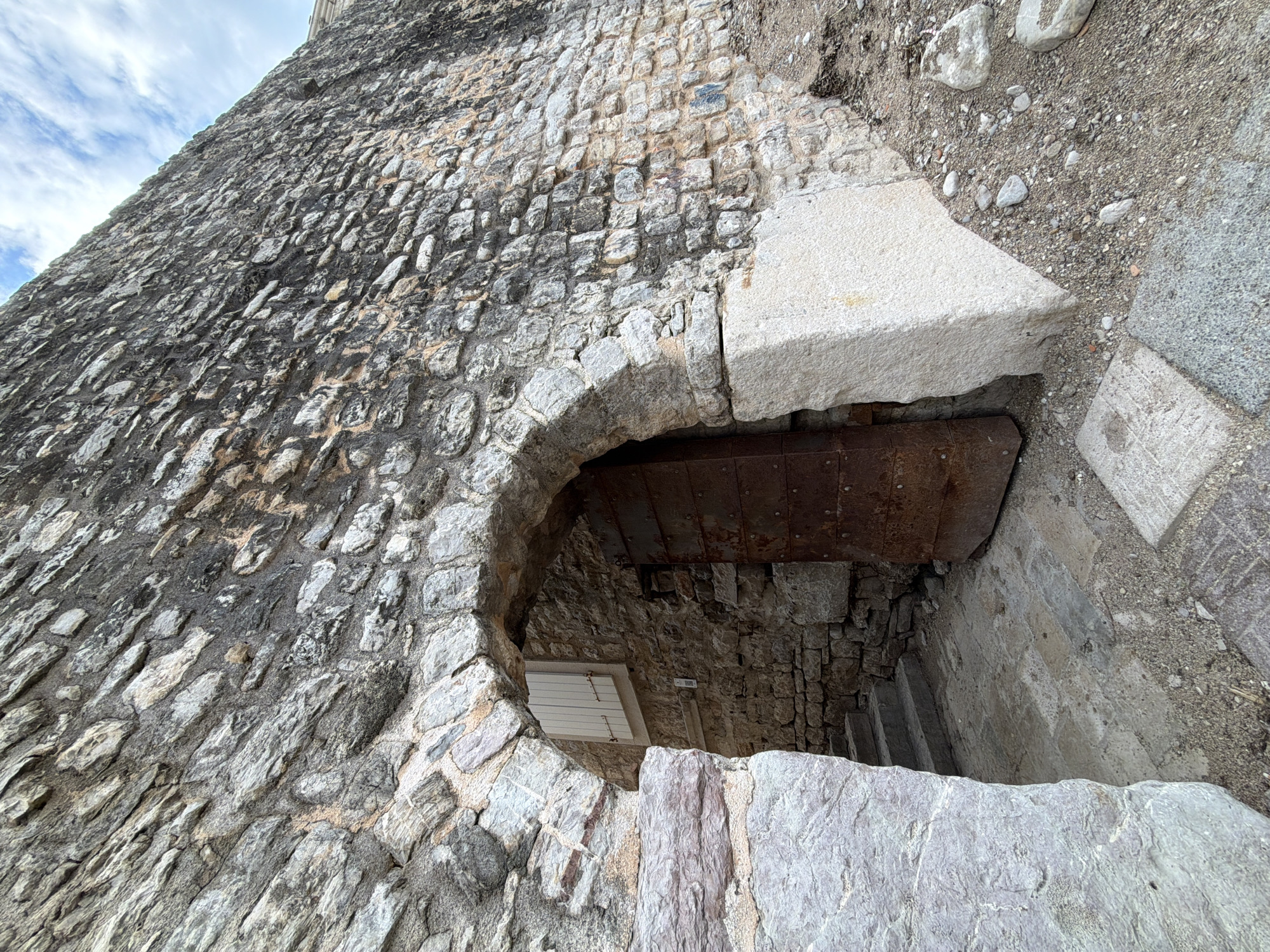 The image shows a close-up view of a section of a stone structure, possibly a tower or building, located in Budva, Montenegro. The structure is characterized by its rough, irregularly shaped stones, which form a wall that is partly weathered.
A prominent feature is an arched opening, which reveals a dark interior with an internal wooden or metal beam. Inside the opening, a small, white, possibly wooden door is visible.
The ground around the structure is covered with gravel and stone debris. The overall impression is of an old, historical construction, possibly a fortification or part of an old town wall. The environment suggests a historical or tourist site in Budva, Montenegro.
