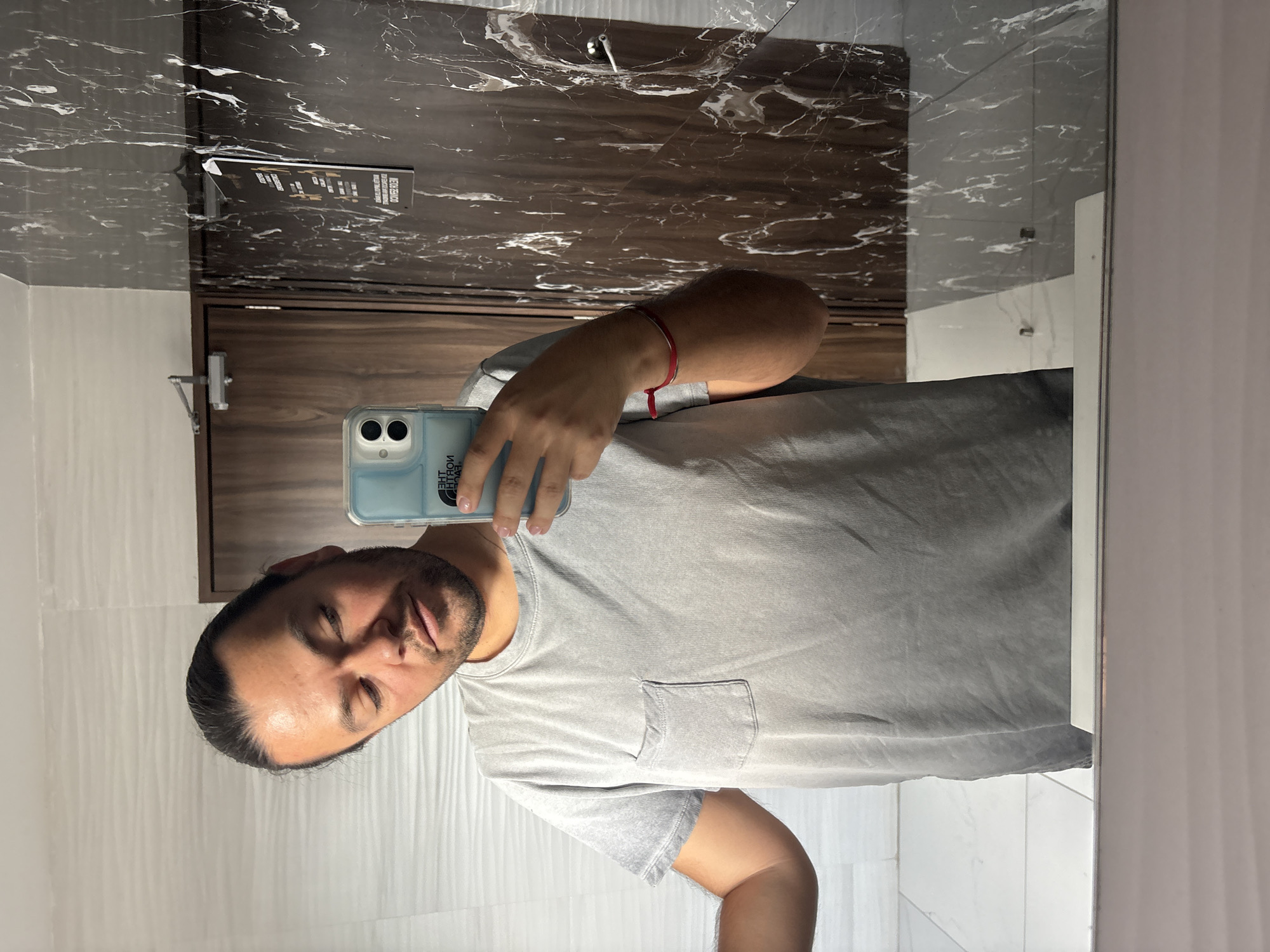 A man takes a selfie in front of a mirror in a bathroom. The mirror reflects a wall with brown, marbled tile. The man wears a grey t-shirt with a pocket and a red bracelet on his left wrist. He holds a light blue smartphone. The environment consists of white tiles, a door, and a sign. There are no details indicating the location is Puebla, Mexico.