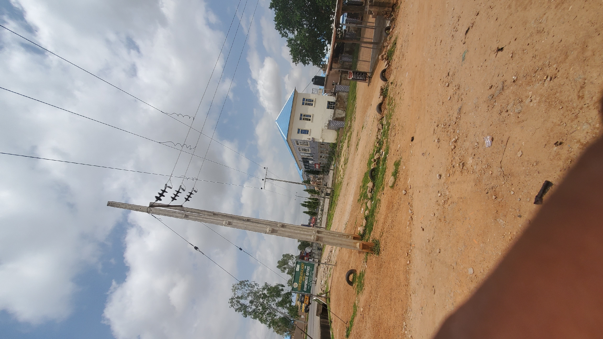 The image depicts a street scene in Jalingo, Nigeria. In the foreground, a dusty, reddish-brown dirt road is visible, littered with small stones and debris. Two discarded car tires lie on the ground near the edge of the road. A wooden utility pole with insulators and overhead wires dominates the left side of the frame. To the left of the pole, a sign is mounted on a metal pole. The sign is green and white and reads "NIGERIAN METEOROLOGICAL AGENCY (NIMET) CORPORATE OFFICE". Behind the sign, a sparse tree with light green foliage is present.
In the middle ground, a two-story building with a light blue roof and white exterior walls stands prominently. The building has multiple windows with blue frames and white shutters. A black water tank is visible on top of a smaller structure next to the main building. A rudimentary open-sided shelter with a corrugated metal roof is situated to the right of the building, with some items stored inside and a sign that reads "HOTEL". A fence made of concrete blocks and metal bars encloses a portion of the property.
In the background, more trees and a cloudy blue sky are visible. A yellow bus is parked on the left side of the road, further in the distance. Several other trees line the street in the middle distance, and a few smaller utility poles are also scattered throughout the scene. The overall environment appears to be a mix of urban infrastructure and undeveloped land.