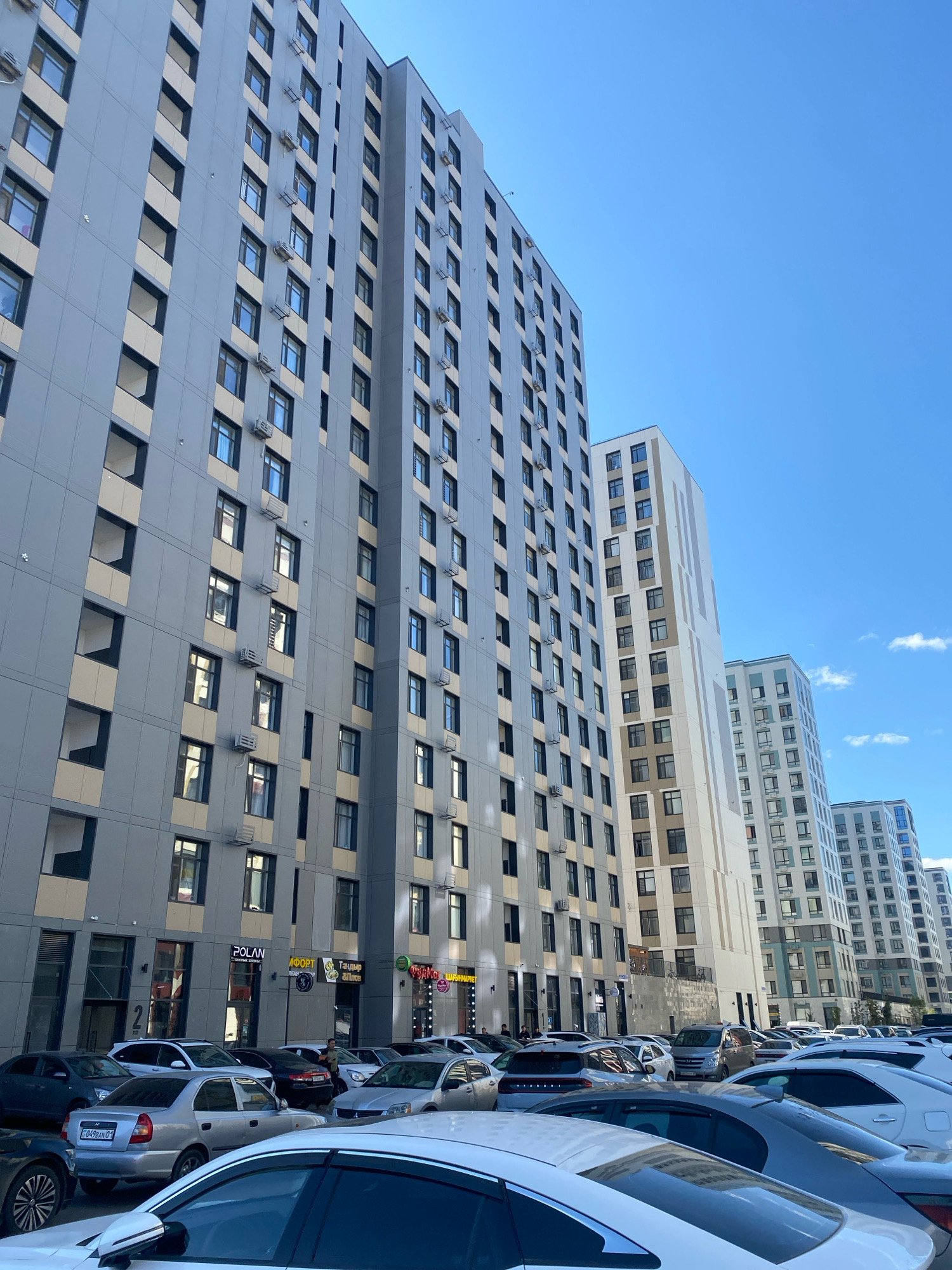 The image depicts a modern urban scene in Astana, Kazakhstan, dominated by several high-rise residential buildings with a predominantly gray and beige color scheme. The buildings feature numerous windows and are equipped with exterior air conditioning units. In the foreground, a line of parked cars is visible, including a silver Hyundai Accent with license plate C049EAN 01, and several other sedans and SUVs. The sky is a clear blue with a few wispy clouds. Some storefronts are visible at the base of the buildings, with illuminated signs for businesses such as "POLAN" and "АФФОРТ". People are present, with some walking along the sidewalk and others near the parked cars. The overall impression is of a bustling residential and commercial area.