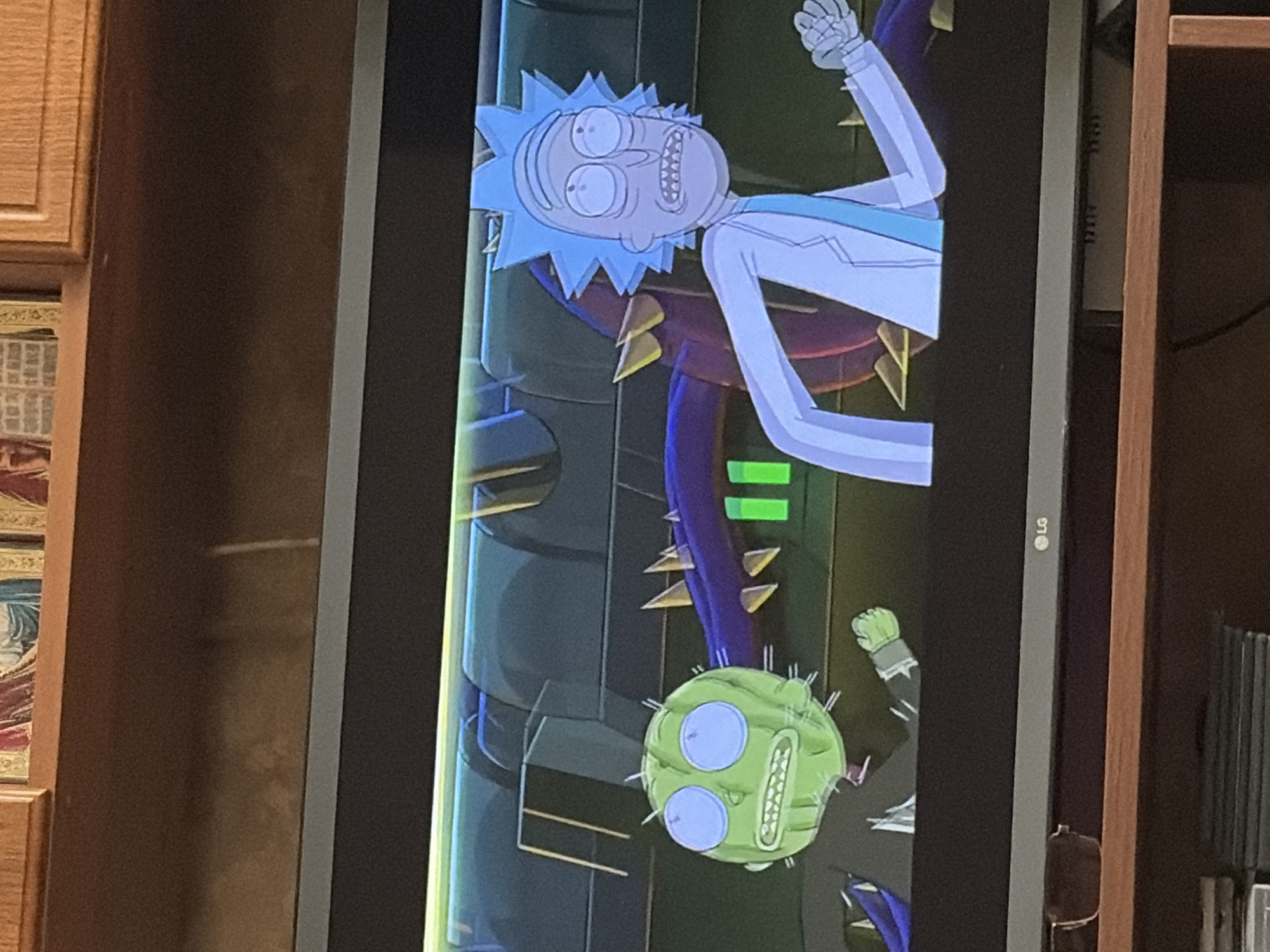 An animated scene from the television show Rick and Morty is displayed on a television screen. The scene features Rick Sanchez and a character that appears to be a watermelon-like creature with spikes, both in mid-action. Rick is wearing his signature white lab coat and blue shirt, with his arms outstretched as if running or in motion. The watermelon creature, with large blue eyes and sharp teeth, is also depicted in a running pose, its spiky body visible. Behind them, a purple, spiky tentacle-like object extends across the frame, and a blurred, metallic background suggests an industrial or alien environment. The television screen itself is placed on a wooden surface, with a dark bezel surrounding the display. The ambient lighting in the room is dim, with the light from the television illuminating the immediate foreground.