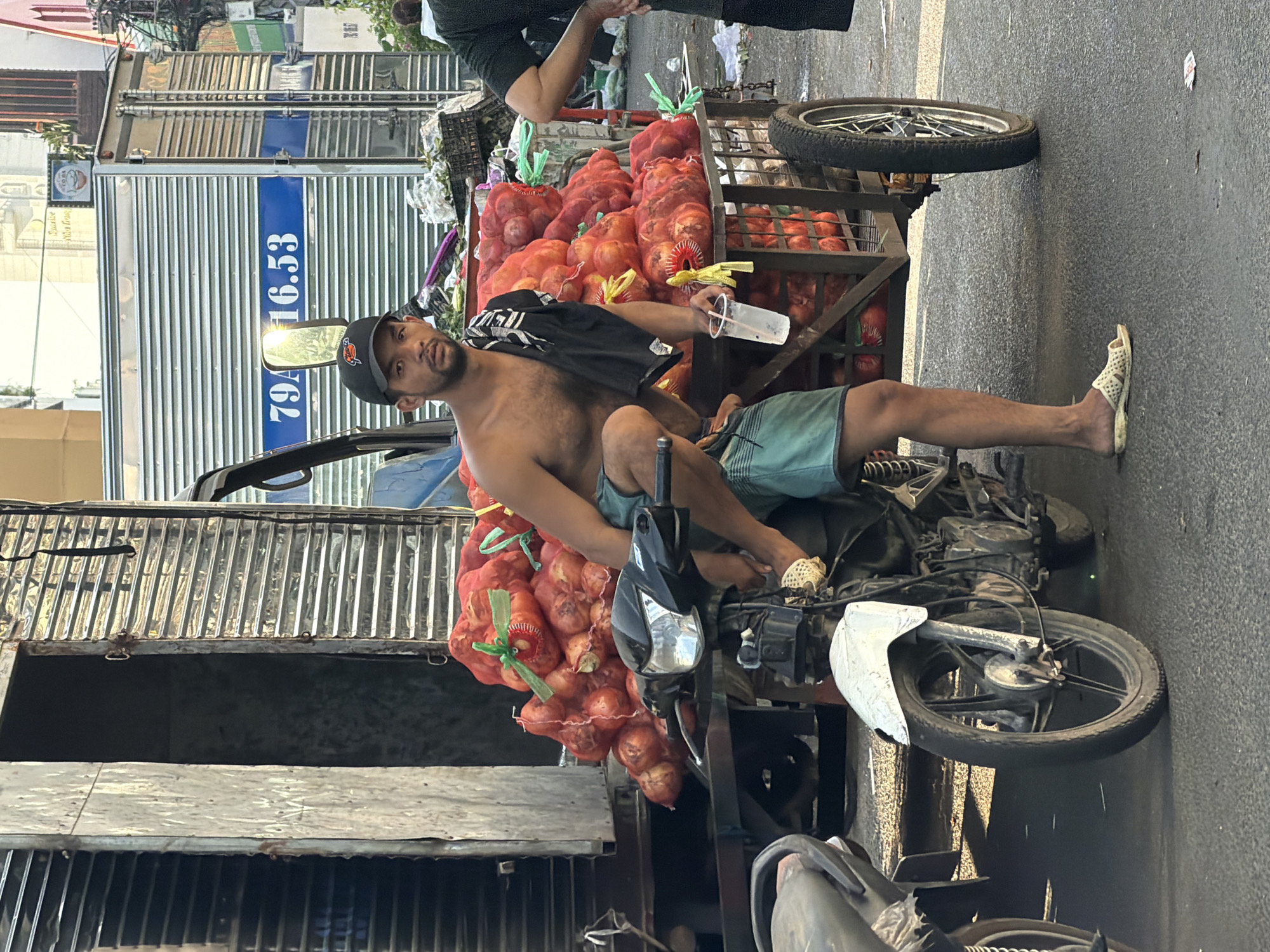 A shirtless man wearing a black baseball cap and turquoise shorts squats on a motorcycle, holding a plastic cup with a beverage. He is next to a cart loaded with large nets of red onions, tied with green netting. The motorcycle has a white front fender and a black body. In the background, a large truck with blue lettering and the number "79A.16.53" is visible. The scene appears to be a street market in Nha Trang, Vietnam, with other market stalls and buildings in the distance. The lighting suggests it is daytime, possibly late afternoon, given the shadows.