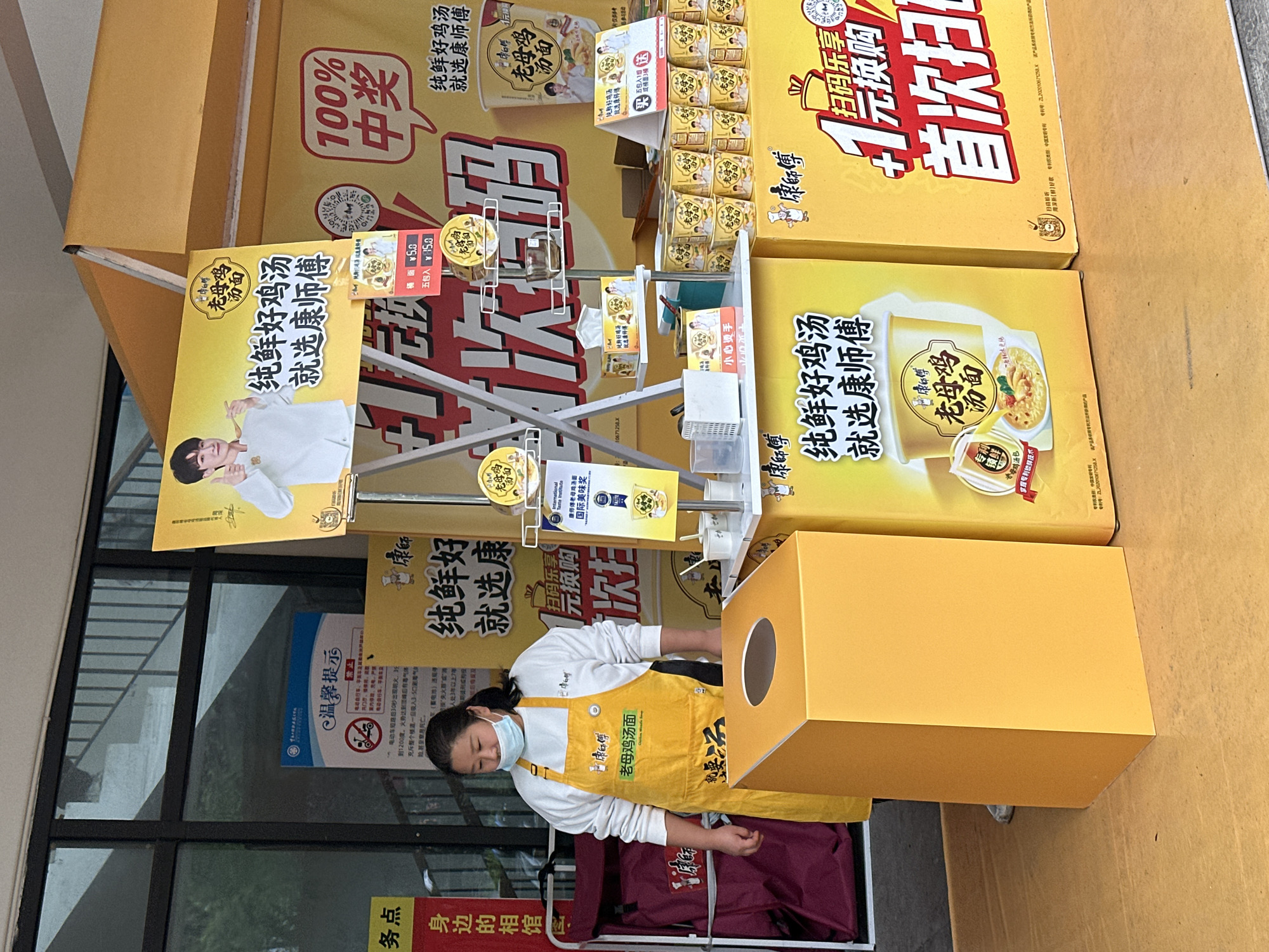 The image shows a woman standing behind a promotional display for "Kang Shi Fu" chicken noodle soup, likely located in Jiangjin, China. The display consists of several yellow cardboard boxes with the product advertised in Chinese. The boxes contain instant noodle cups and multi-packs. The display also features posters with images of a man, presumably a celebrity endorser, and the product itself. The woman is wearing a yellow apron with Chinese writing on it and a face mask. She appears to be preparing something within a yellow cardboard structure, which contains a hole on the upper right side, in front of the display. The image is taken outdoors, near a window, with some greenery reflected in it. A red folding chair is behind the woman.