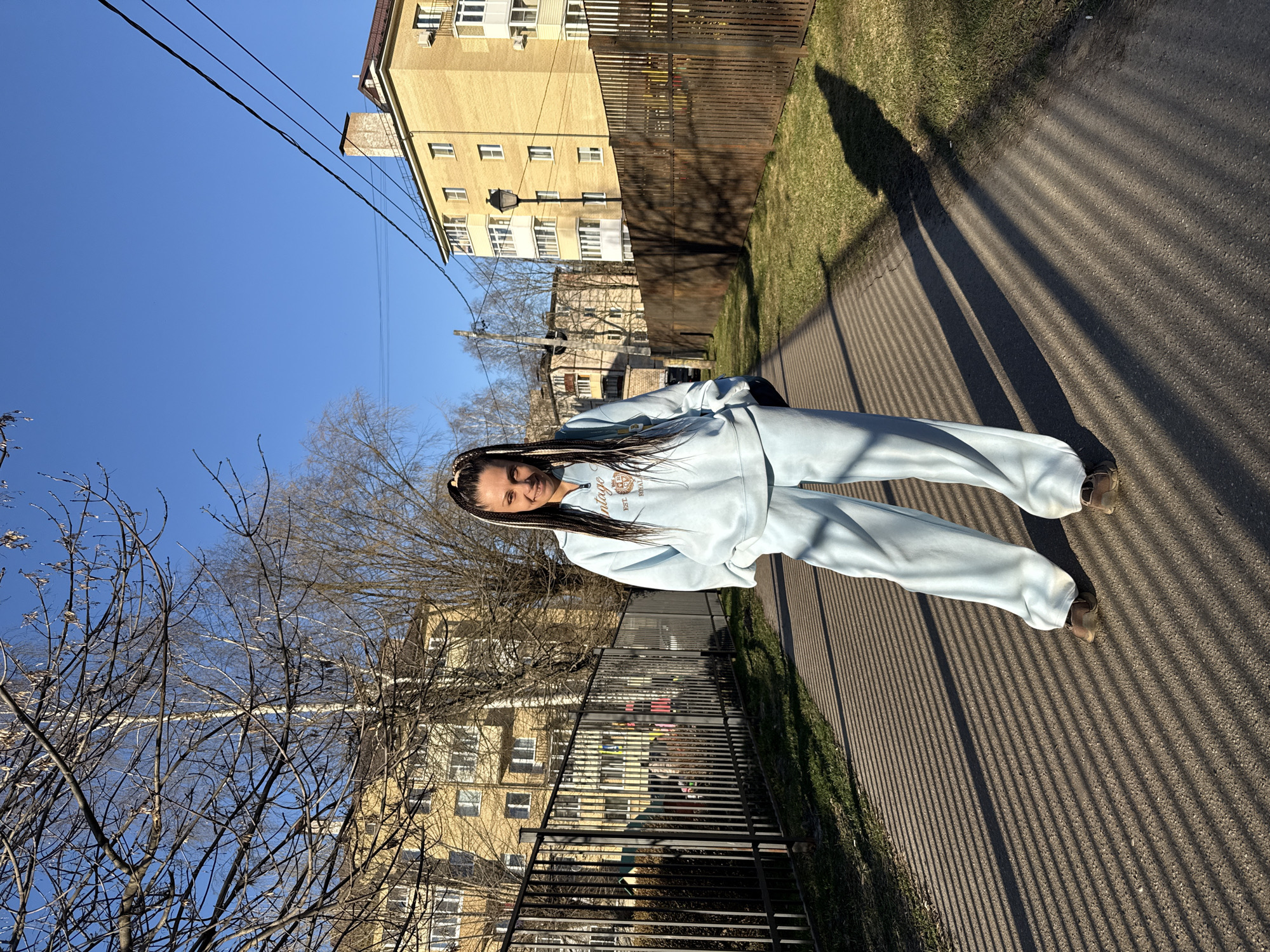 A young woman with long, braided hair stands on a paved path in a residential area of Rusanovo, Russia, on a clear, sunny day. She is wearing a light blue tracksuit, featuring a sweatshirt with the text "VINTAGE" and "EST. ROYAL" visible on it. Her hands are in her pockets, and she exhibits a relaxed, slight smile.
The scene is bathed in bright sunlight, casting long, distinct shadows from a black metal fence onto the asphalt path. These shadows run diagonally across the foreground and midground, indicating either early morning or late afternoon. To the right of the path is a strip of green grass.
In the background, several buildings are visible. A prominent yellow brick apartment building with white balconies stands behind a dark wooden fence. Further back, another, older-looking building can be seen through the bare branches of trees, suggesting late winter or early spring. Overhead utility wires traverse the bright blue sky on the left side of the frame. A streetlamp is also visible in the mid-distance. The overall atmosphere is bright and serene, capturing a moment of casual outdoor presence in an urban setting.