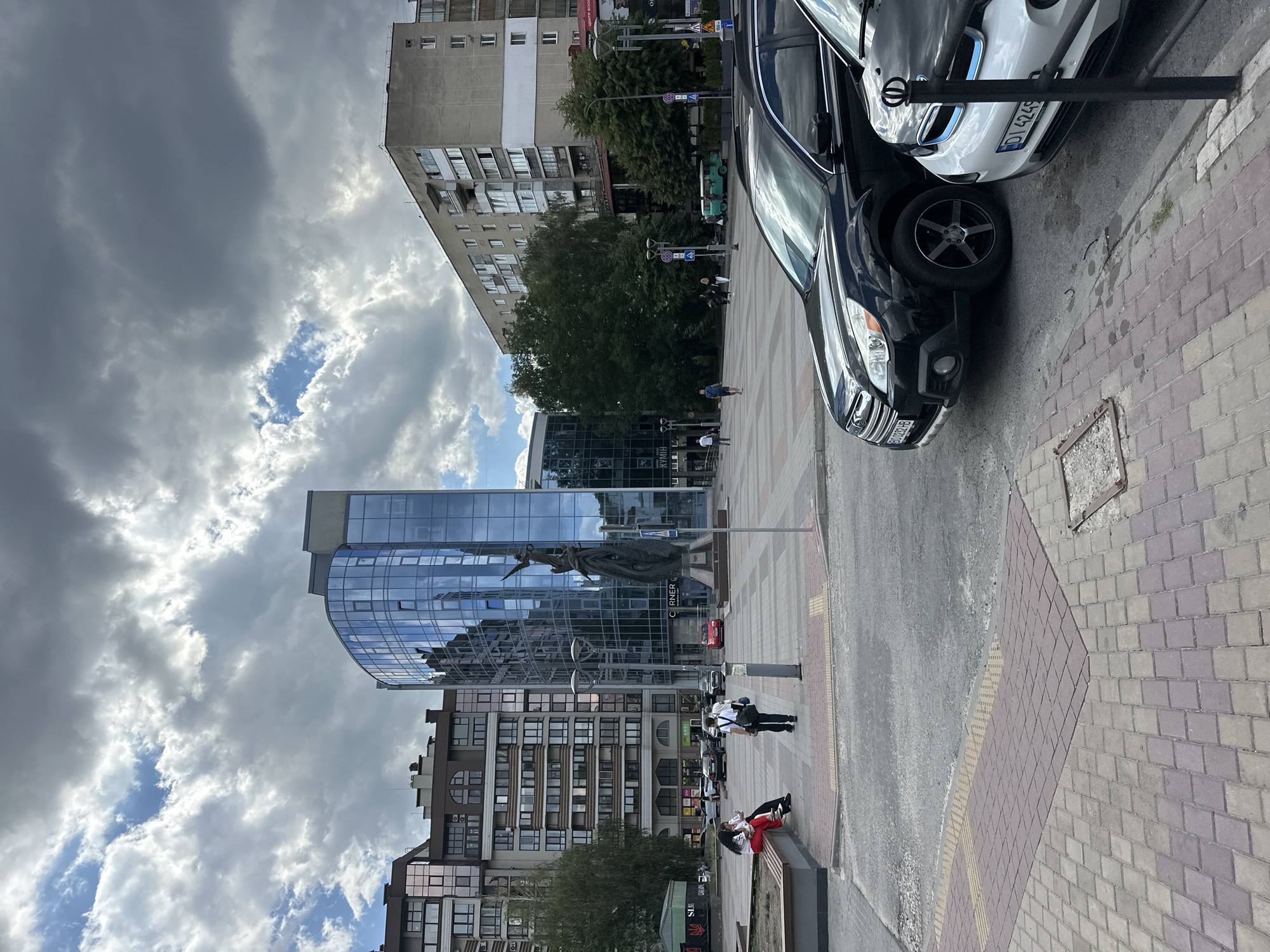This is an outdoor, eye-level, vertical, medium shot of a city square in Ternopil, Ukraine, on a cloudy day. In the foreground, two cars are parked on the right side of the frame: a black Subaru Outback and a white BMW i3. The Subaru's license plate reads B0 0282 HB. The BMW's license plate reads DI 4249.
In the middle ground, a paved square is visible with several people walking. To the left, a long bench is occupied by two individuals wearing red and white attire. A third person, wearing a white shirt and black pants, walks away from the bench towards the right. In the center of the square, a tall, dark statue stands in front of a modern, curved glass building. The building has "CORNER" visible in white lettering. Adjacent to the glass building is a multi-story residential building with numerous windows and balconies.
In the background, more buildings are visible, including a tall, grey, Soviet-era apartment block on the right. Trees are scattered throughout the square and between buildings. The sky is filled with heavy, grey clouds, with patches of blue sky visible.