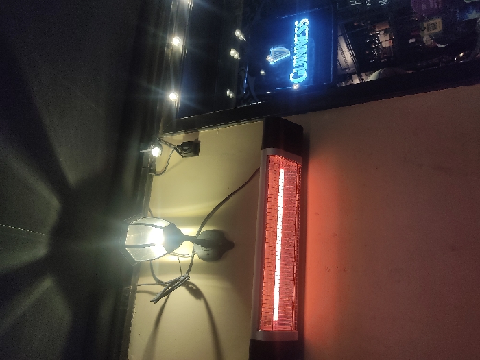 The image depicts an outdoor seating area, likely a patio or terrace, possibly in Moscow, Russia, due to the presence of a Guinness advertisement sign. A wall forms the backdrop, and attached to it are an outdoor lamp and an electric heater emitting a red glow. The lamp casts a bright light and a distinct shadow on the adjacent wall. String lights hang above. The wall is painted a light color. The setting suggests a nighttime or dimly lit environment. The heater and lighting indicate the space is set up for use in cooler weather.
