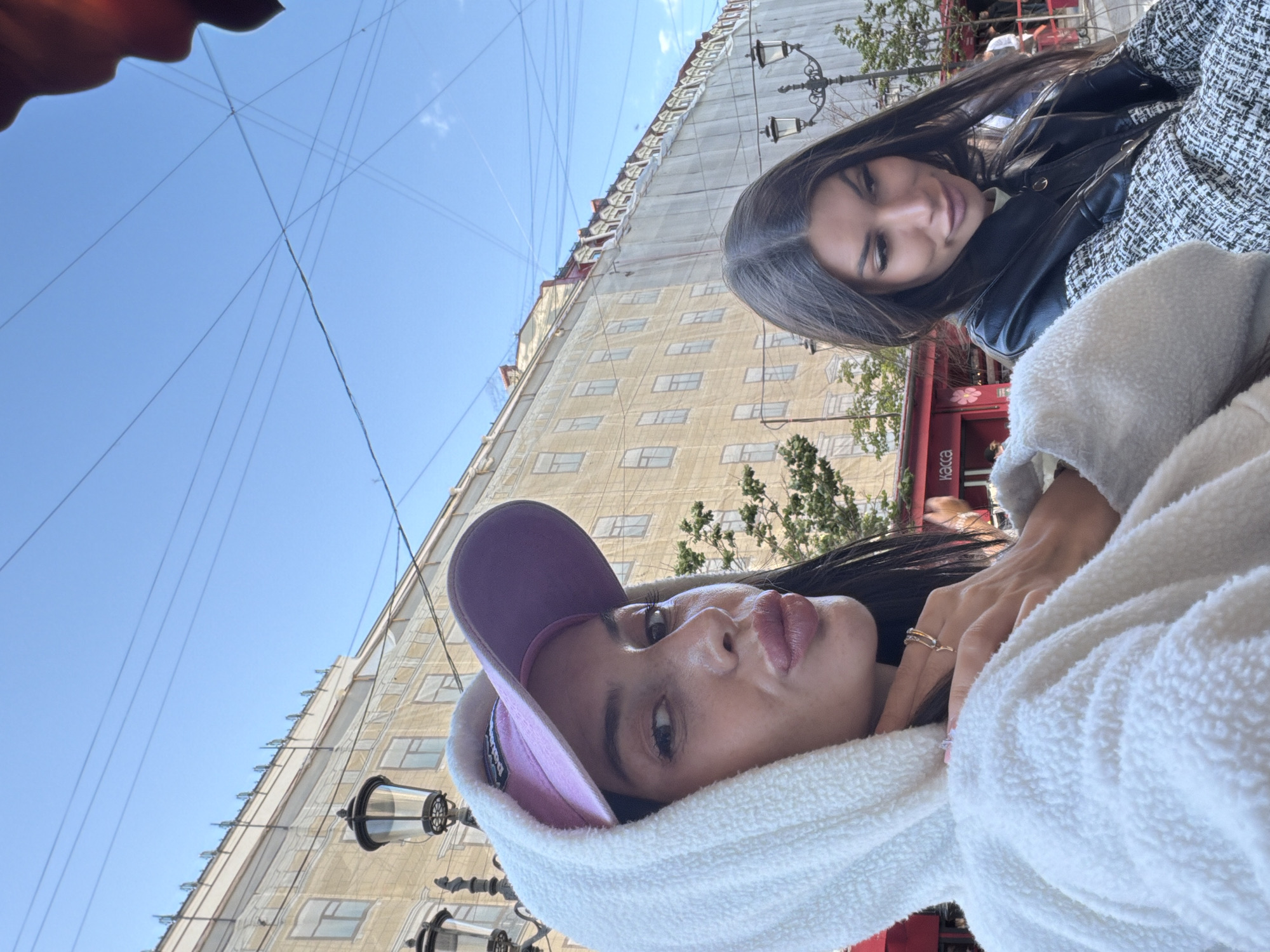 Two young women are captured in a close-up, outdoor selfie on a sunny day in front of a building draped in scaffolding or protective sheeting. The woman in the foreground, wearing a white hooded fleece and a purple baseball cap, looks directly at the camera with her lips pursed. She has dark hair and is wearing makeup. Her hand, adorned with a ring, is resting on her neck. To her right, the second woman, who appears to be her twin, is looking down and to the side. She has long dark hair and is wearing a black leather jacket over a patterned shirt. She is also wearing makeup. The background features a building with multiple windows, streetlights, and a clear blue sky with visible overhead wires. A red structure with the word "КАССА" (KASSA - meaning "cash desk" or "ticket office" in Russian) is visible behind the women, suggesting a commercial area.
