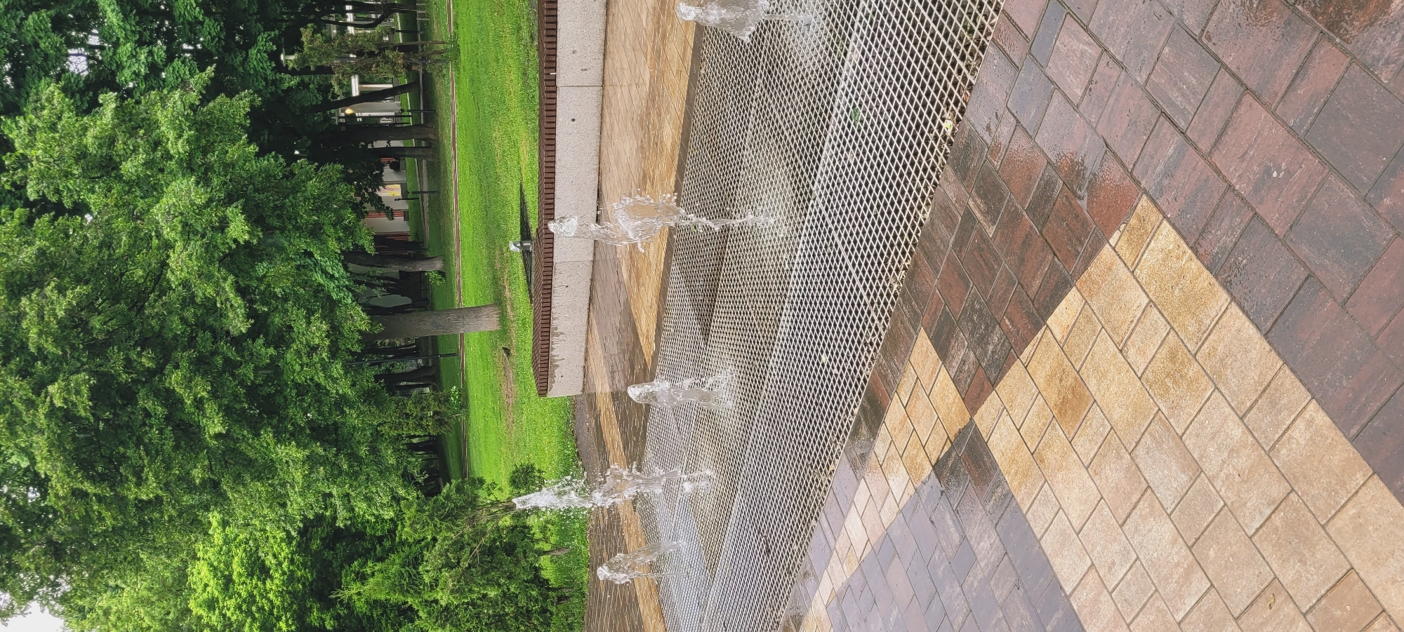 The image depicts a park scene in an unknown city. The focus is on a ground-level fountain with water jets spraying upwards through a metal grate. The fountain's perimeter is defined by brown and tan brickwork. Adjacent to the fountain is a well-manicured green lawn, bordered by trees. The background includes a building visible through the trees.