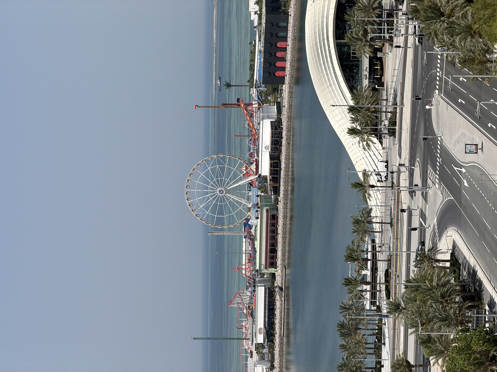 An aerial, eye-level, long shot captures a waterfront amusement park on a clear, sunny day. The water, a tranquil blue, meets the horizon where it blends with a pale blue sky. In the mid-ground, the amusement park features a prominent white Ferris wheel and a red roller coaster, surrounded by various smaller rides and structures. Buildings with dark roofs and light-colored walls are situated along the waterfront, some with large arching windows. A modern, white, wave-shaped building with glass panels dominates the foreground, with a road curving around it. Palm trees are scattered throughout the scene, lining the streets and the waterfront. Several cars are visible on the road, and a small boat is present in the distant water. The scene is devoid of people.