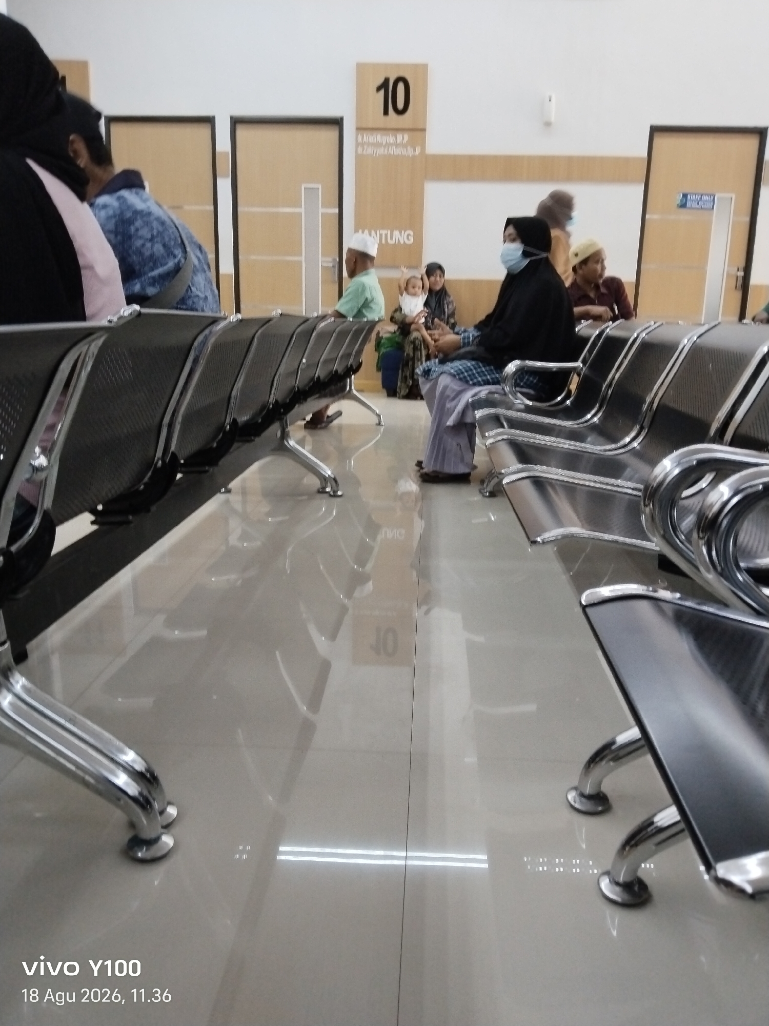 The image depicts a waiting area, likely in a clinic or hospital in Patokan, Indonesia. In the foreground, a row of dark, metal bench seating with chrome legs is visible, reflecting on the polished tiled floor. Several people are seated in the chairs, including a woman wearing a black hijab and a face mask, and a child being held by another person. Further back, two wooden doors are visible, one with a sign indicating "10" and another with a "STAFF ONLY" sign. The lighting appears to be from overhead fluorescent fixtures, casting reflections on the floor. The overall atmosphere suggests a quiet, institutional setting.