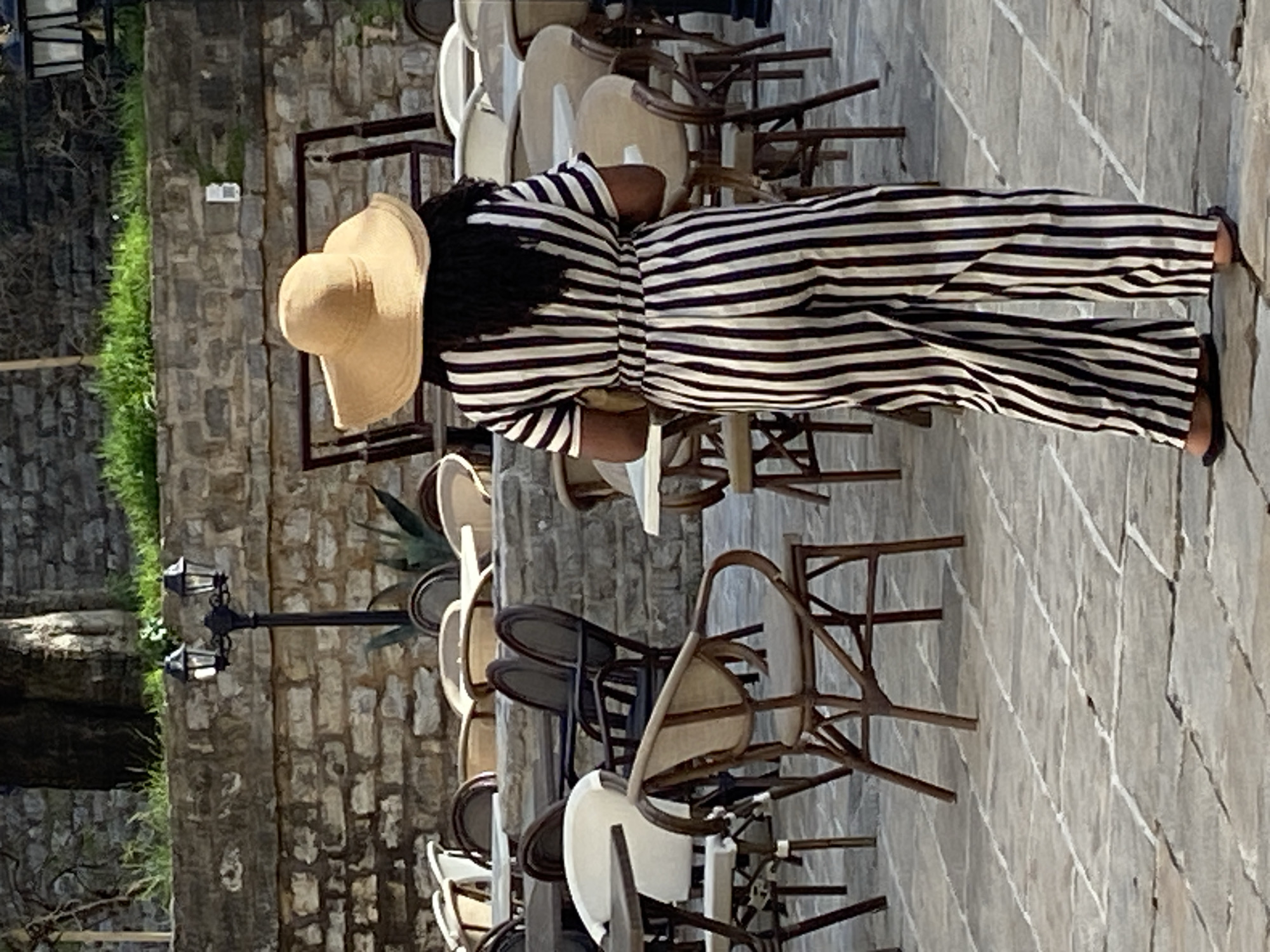 A woman in a striped jumpsuit and a large straw hat stands on a stone-paved surface. She is surrounded by stacked chairs, suggesting a cafe or restaurant setting. A stone wall is to the left with some greenery. A black lamppost is also visible. This image appears to be taken in an old European city, possibly Budva, Montenegro.
