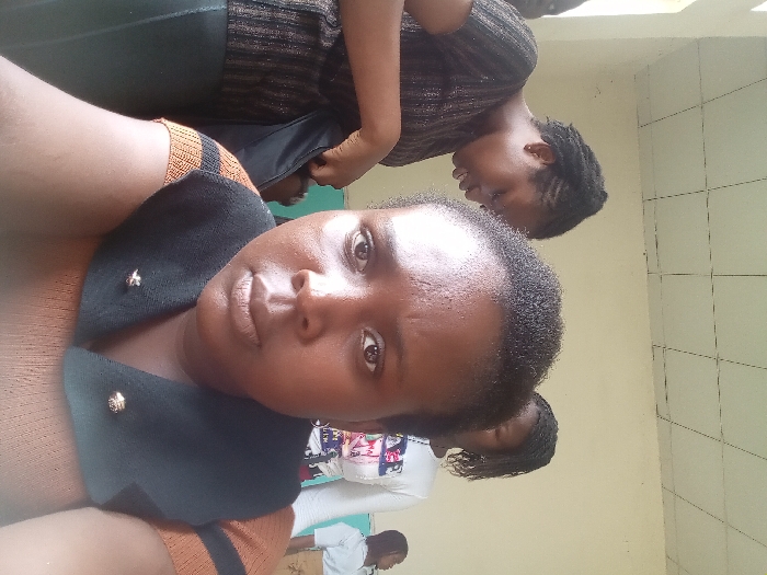 An indoor close-up photograph taken in OGBOKO, Nigeria, depicts four individuals. The primary subject in the immediate foreground is an adult female with dark skin, short dark hair, and a neutral expression, gazing directly at the camera. She wears an orange-brown short-sleeved top with black horizontal stripes, layered with a black V-neck vest featuring a pointed collar, secured by two small, gold-colored brooches, possibly shaped like bees.
Behind and to the left of the foreground subject, a second adult female with dark braided hair is visible, facing slightly away from the camera and looking downwards. She wears a dark, possibly striped, short-sleeved top and appears to be interacting with a dark item, potentially a bag or garment, at her waist.
Partially obscured by the foreground subject's head, a third adult female with dark locs stands in the mid-background. She wears a white short-sleeved top adorned with a multi-colored graphic design on the chest. A fourth individual, wearing a white top, is partially visible on the far right side of the frame.
The environment features light-colored, plain walls and a white ceiling with a visible grid pattern, indicative of ceiling tiles.