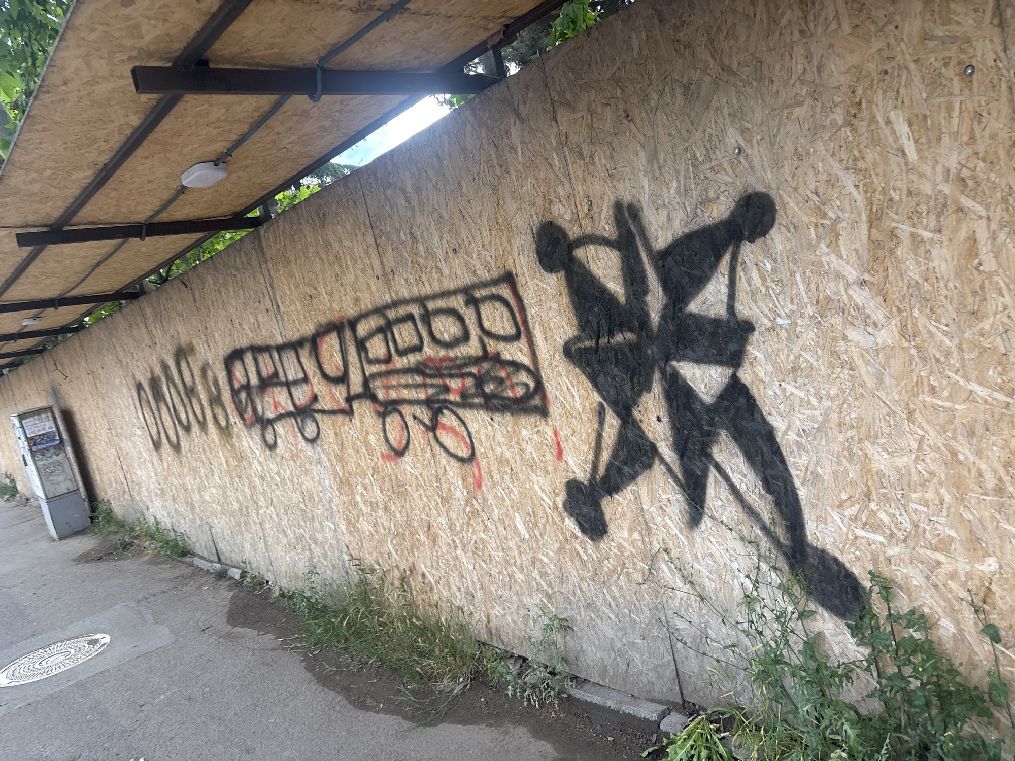 The scene depicts a weathered wooden wall alongside a paved walkway in Tbilisi, Georgia. The wall is covered in graffiti, featuring a crudely drawn bus or tram in black and red, indiscernible writing, and a black abstract design reminiscent of a complex snowflake or a stylized starburst. A partial roof structure extends over the wall, consisting of a wooden surface supported by metal beams, with an overhead light fixture visible. Weeds and grass grow along the base of the wall. A utility box is visible in the background. The overall environment suggests a semi-urban setting.