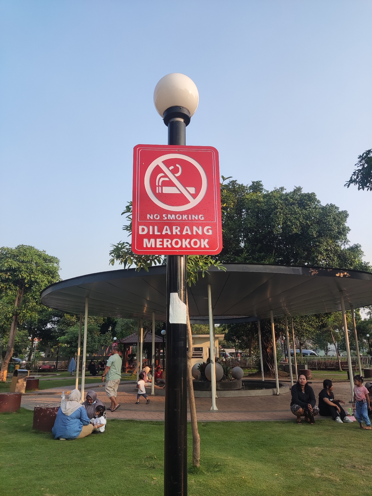 The image depicts a public park setting in Pagerwojo, Indonesia, under a clear blue sky. A prominent red sign with a white no-smoking symbol and the text "NO SMOKING DILARANG MEROKOK" is mounted on a lamppost in the foreground. The lamppost is black with white tape near its base and topped with a white globe light.
To the left of the lamppost, a woman in a blue hijab and a long-sleeved blue shirt sits on the grass with two children. One child is wearing a white hijab and a pink dress, and the other is wearing a white shirt and brown pants. They are facing towards the right of the frame.
In the middle ground, a covered pavilion with a dark roof supported by white pillars is visible. Several people are seated or standing within and around the pavilion. A man in a light green shirt and beige shorts walks away from the camera. Another person, possibly a child, wearing a white shirt and blue shorts runs towards the right.
To the right of the pavilion, a group of people are seated on the grass. Two women are squatting, one wearing a black long-sleeved shirt and patterned skirt, and the other wearing a black shirt and dark pants. A young girl in a pink t-shirt and blue jeans stands near them.
The park features well-maintained green grass, paved pathways, benches, and lush trees. In the background, a few buildings and a fence are visible. The overall scene suggests a relaxed afternoon gathering in a community space.