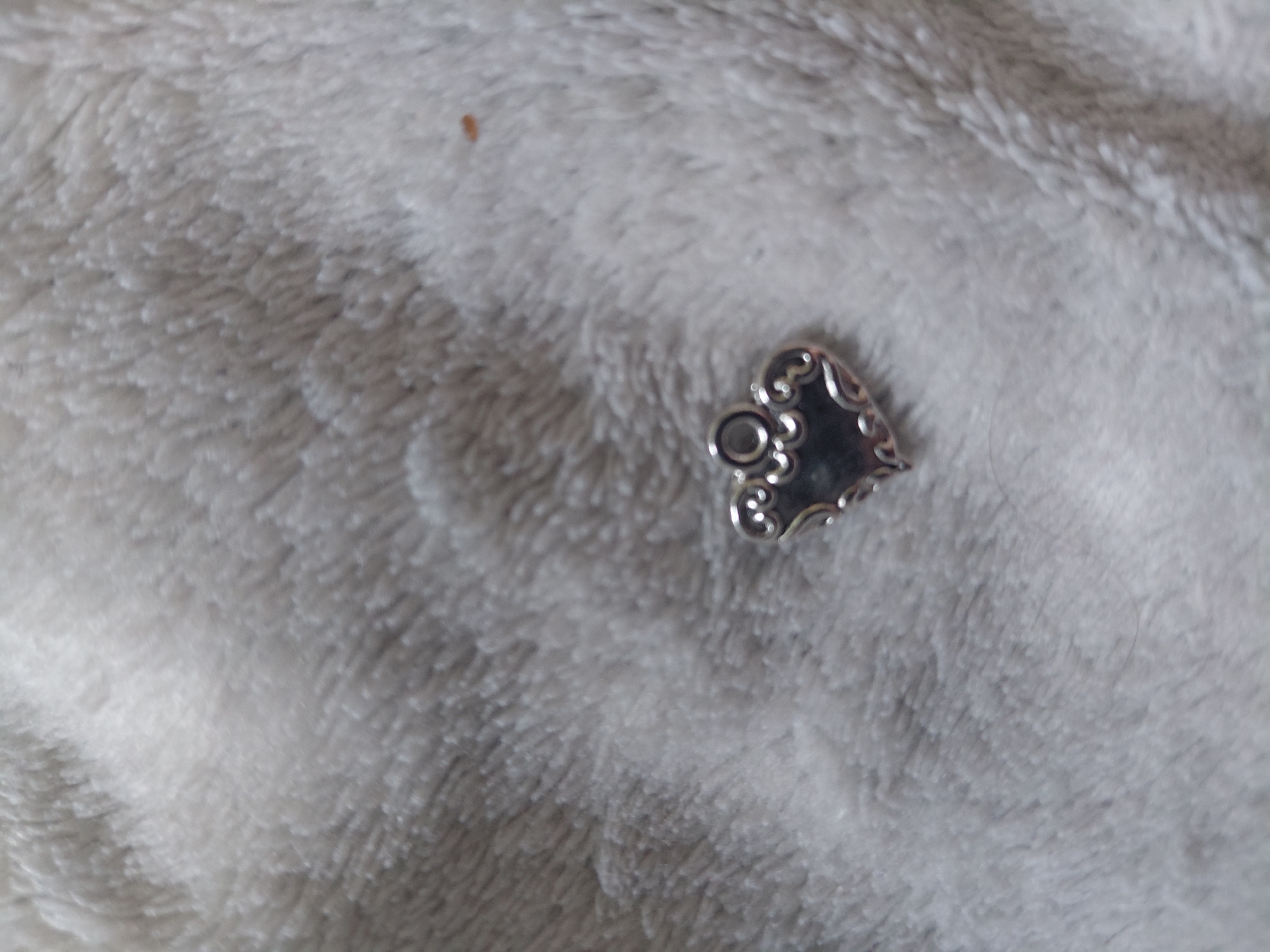 A small, ornate, silver-toned heart-shaped charm, featuring decorative scrolled edges and a circular attachment loop, rests on a textured surface. The surface is composed of soft, light-colored fibers, likely a white or off-white fluffy textile material such as faux fur or a blanket. The scene is located in the city of Kyiv, Ukraine. A singular, minute reddish-brown speck is present on the fabric in the upper right area of the frame, distinct from the charm.