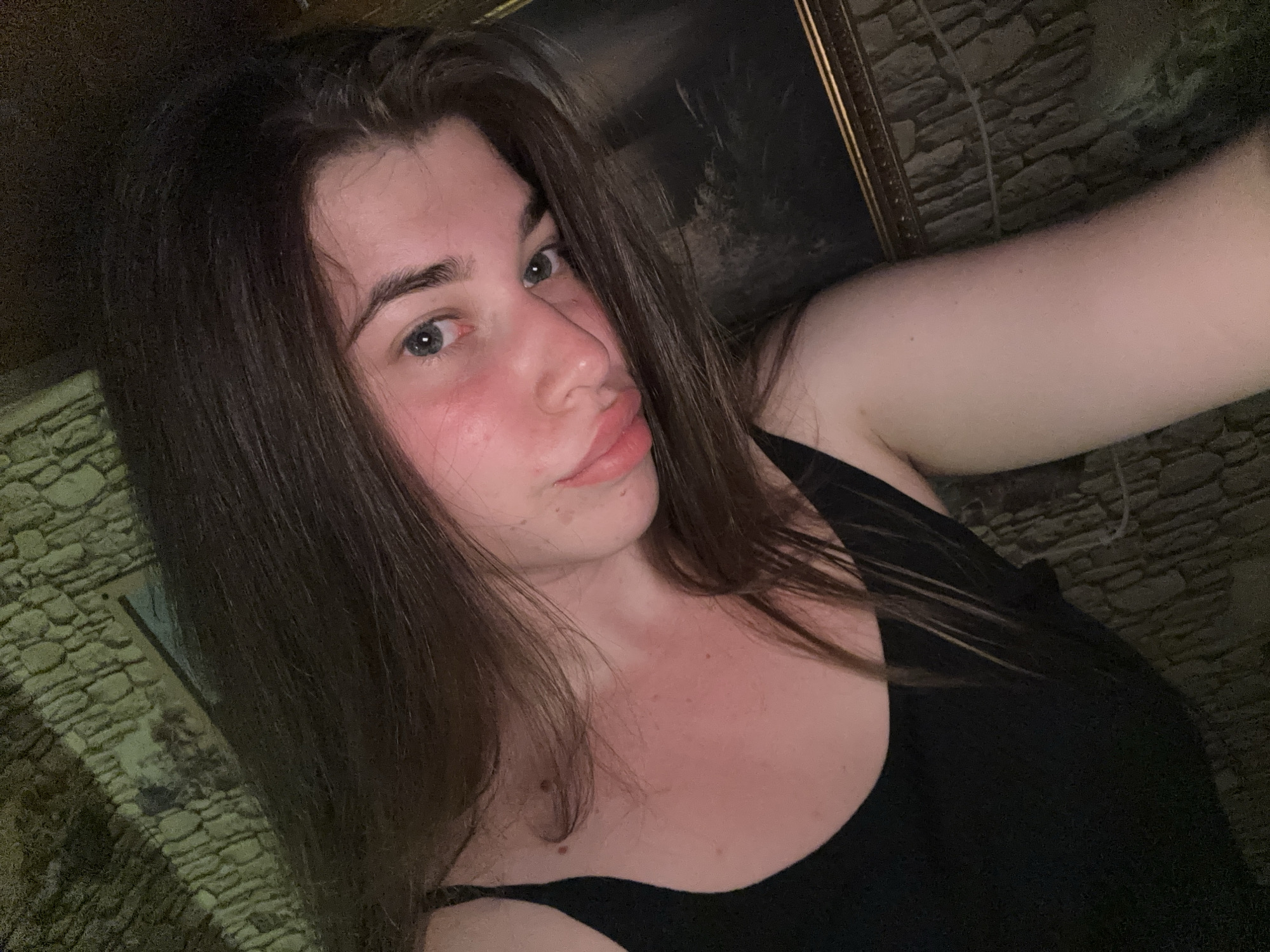 The image is a selfie taken by a young person with dark brown hair, fair skin, and light eyes. The person is wearing a black tank top and is looking towards the camera. The background appears to be a wall made of rough, uneven stones, possibly an exterior wall of a building or a rustic interior. There's a hint of what looks like a framed picture or mirror in the upper right corner. The lighting is dim, casting shadows and highlighting the person's face. The overall setting suggests an indoor or possibly an outdoor evening shot in a location with a rustic architectural style. The location in Мещёры, Russia, is not visually discernible from the image itself; this detail is external information.