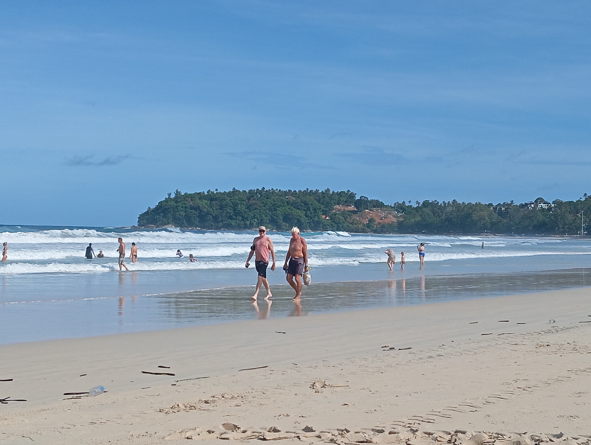 The image depicts a bright, sunny day at Karon Beach in Karon, Thailand. The foreground of the image is a sandy beach with some debris, including sticks and a plastic water bottle. In the middle ground, the ocean meets the shore with gentle waves breaking. Several people are visible in the water and along the shoreline. Two men are walking on the wet sand near the water's edge, one in a pink shirt and dark shorts, the other in dark shorts, carrying a bag. Further out in the water, individuals are swimming and wading. The background features a lush, green headland with trees and some buildings, characteristic of the Karon coastline. The sky is a clear blue with a few wispy clouds.