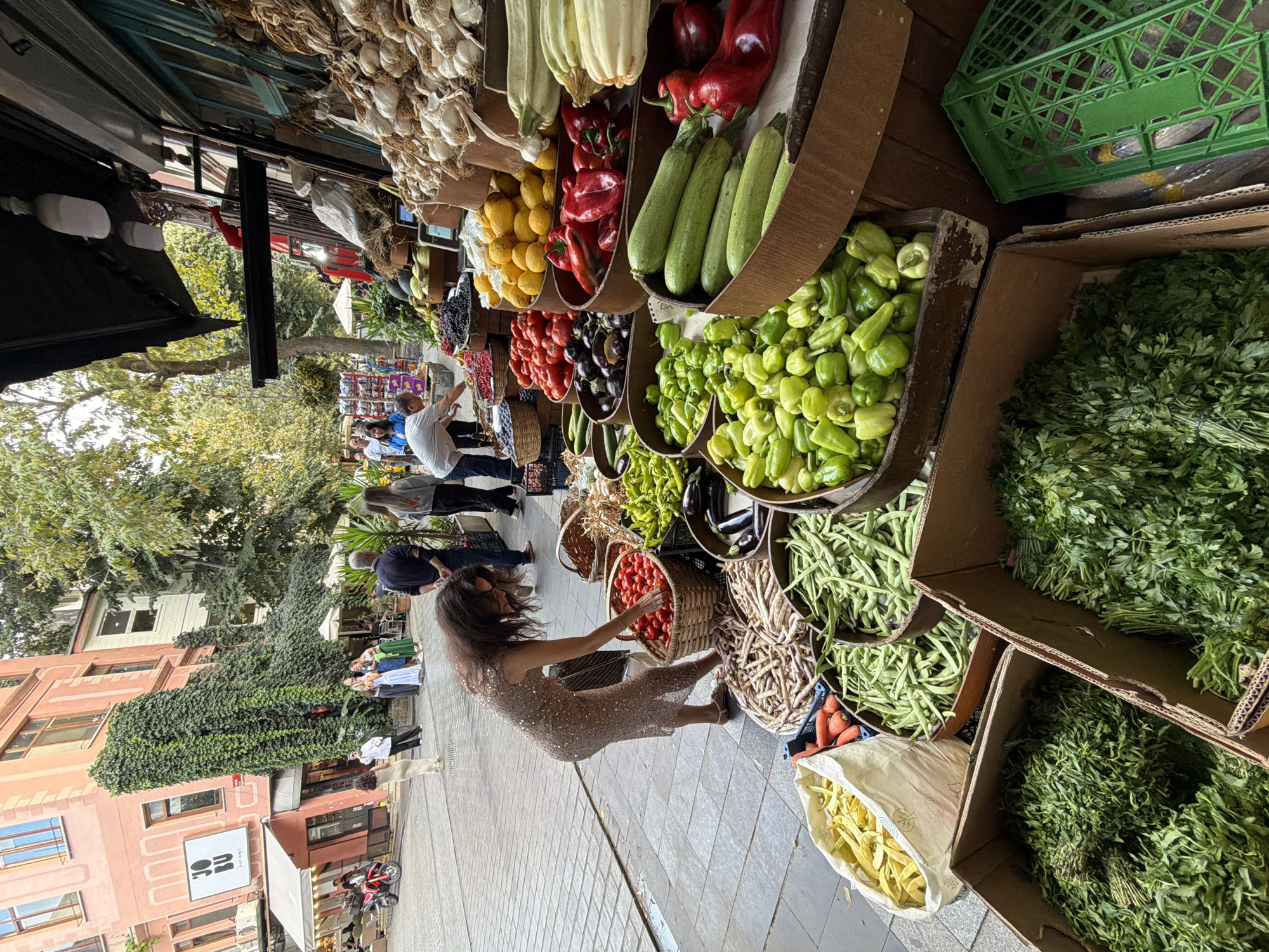 A woman in a sparkly, form-fitting dress is bending down to pick through a basket of tomatoes at an outdoor produce stand on a city street in Üsküdar, Turkey. The street is paved with cobblestones, and a large, pink building with ivy growing up its side looms in the background, adorned with a sign that reads "JOBU". Other shoppers and vendors are visible in the distance. The foreground is dominated by overflowing crates and baskets of fresh vegetables, including green beans, peppers, zucchini, and what appear to be lemons and garlic displayed further back in the stall. The scene is brightly lit, suggesting daytime.