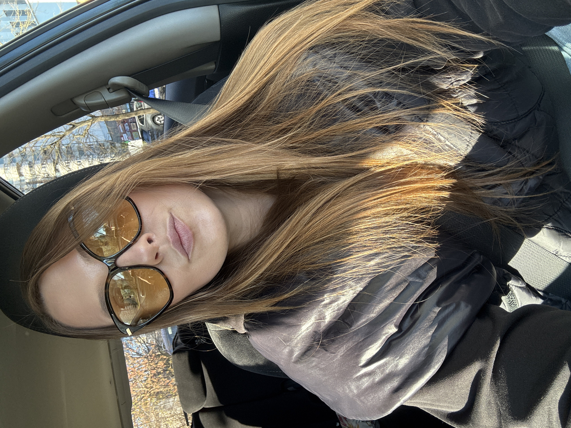 A woman is in the driver's seat of a car, wearing black-rimmed sunglasses with yellow lenses. She has long brown hair. She's wearing a black jacket. The interior of the car is visible, including the seatbelt and part of the dashboard. Through the car window, a street scene with buildings and trees is visible. While the prompt asked for the location in the city of Minsk, Belarus, this cannot be verified based on visual information alone.
