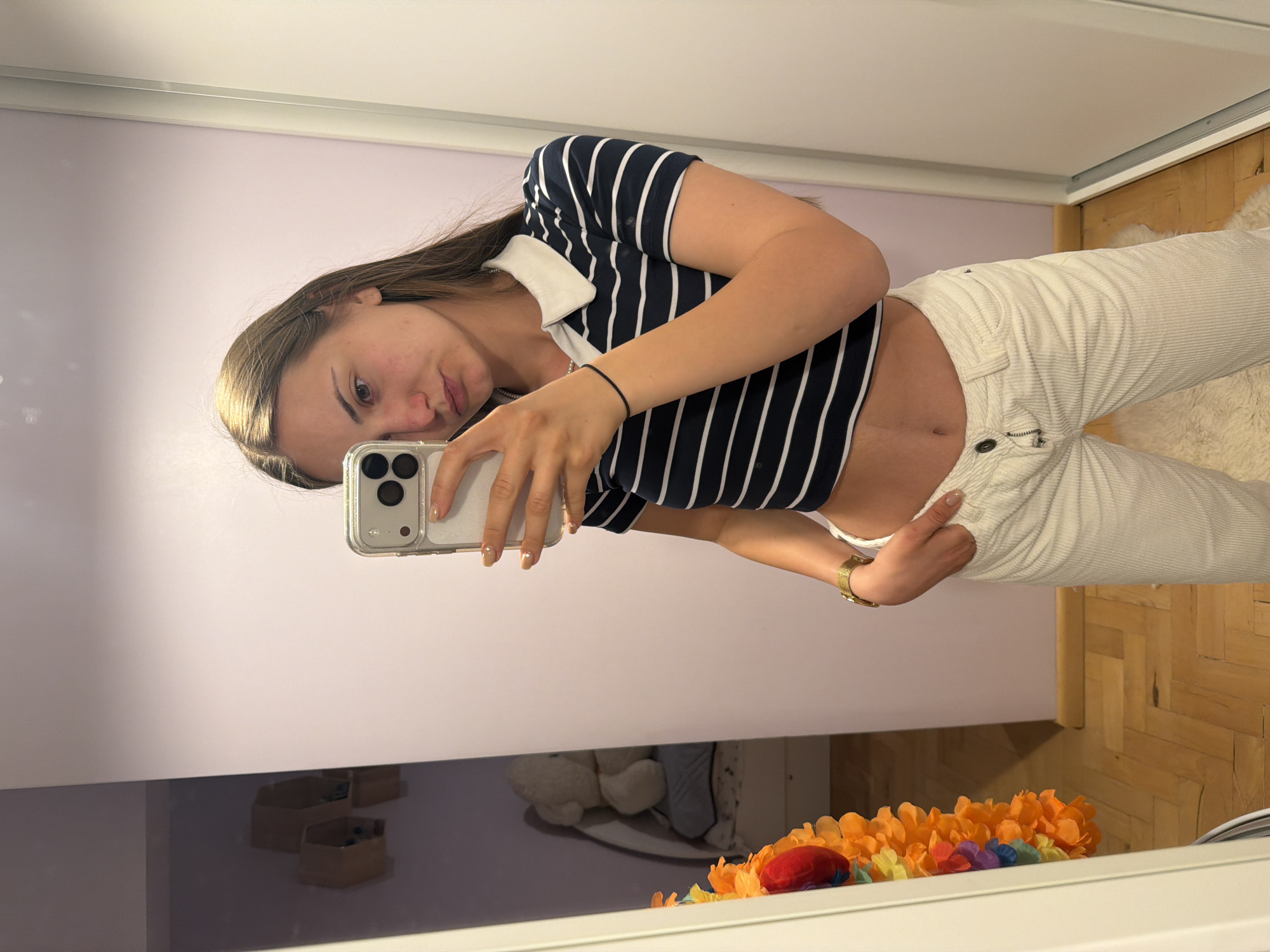 A young woman is reflected in a mirror. She is holding a smartphone in her left hand to take a selfie. She is wearing a navy and white striped polo crop top and white corduroy pants. The background shows a pale lavender wall, a wooden floor, and a white ceiling. A garland of orange and yellow flowers is visible on the mirror's surface. The location cannot be determined from the image.