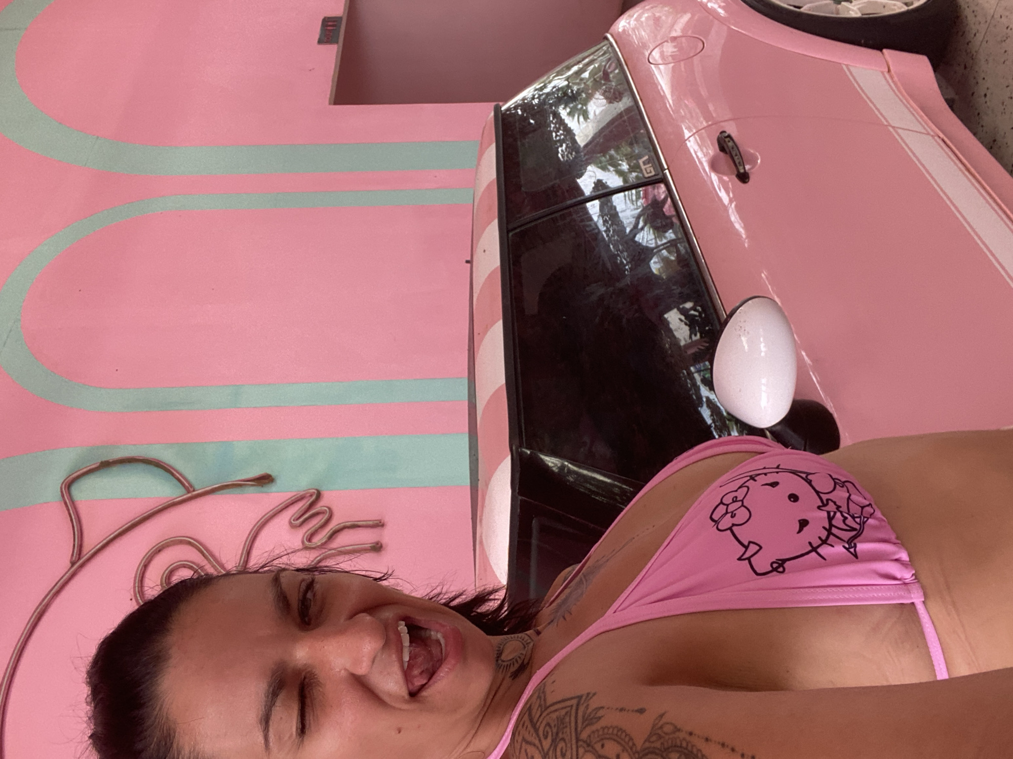 The image captures a playful moment featuring a woman and a pink car, set against a vibrant pink and blue background in what appears to be Tien Thanh Ward, Vietnam.
The primary subject is a woman, visible from the chest up in the foreground. She is looking directly at the camera with a wide, open-mouthed smile and a wink, conveying joy and playfulness. She has dark hair and multiple tattoos, including one resembling an eye on her neck and intricate patterns on her arm and shoulder. She is wearing a pink bikini top featuring a black outline illustration of the character Hello Kitty, adorned with flowers.
Behind the woman, and appearing as if the original photo was taken vertically and then rotated, is a bright pink car with two prominent white racing stripes along its side. The car has dark tinted windows that reflect foliage, suggesting an outdoor or partially open setting. A white side mirror is visible on the car. The letters "GT" can be seen on the rear window of the vehicle.
The background consists of a bold pink wall adorned with thick, light blue, wavy or striped patterns. Near the woman's head, there's an abstract metal art piece with flowing lines integrated into the wall design. The overall scene suggests a themed environment, possibly a cafe, shop, or artistic display, characterized by its highly colorful and coordinated aesthetic. The lighting suggests daytime, possibly in a sheltered area.