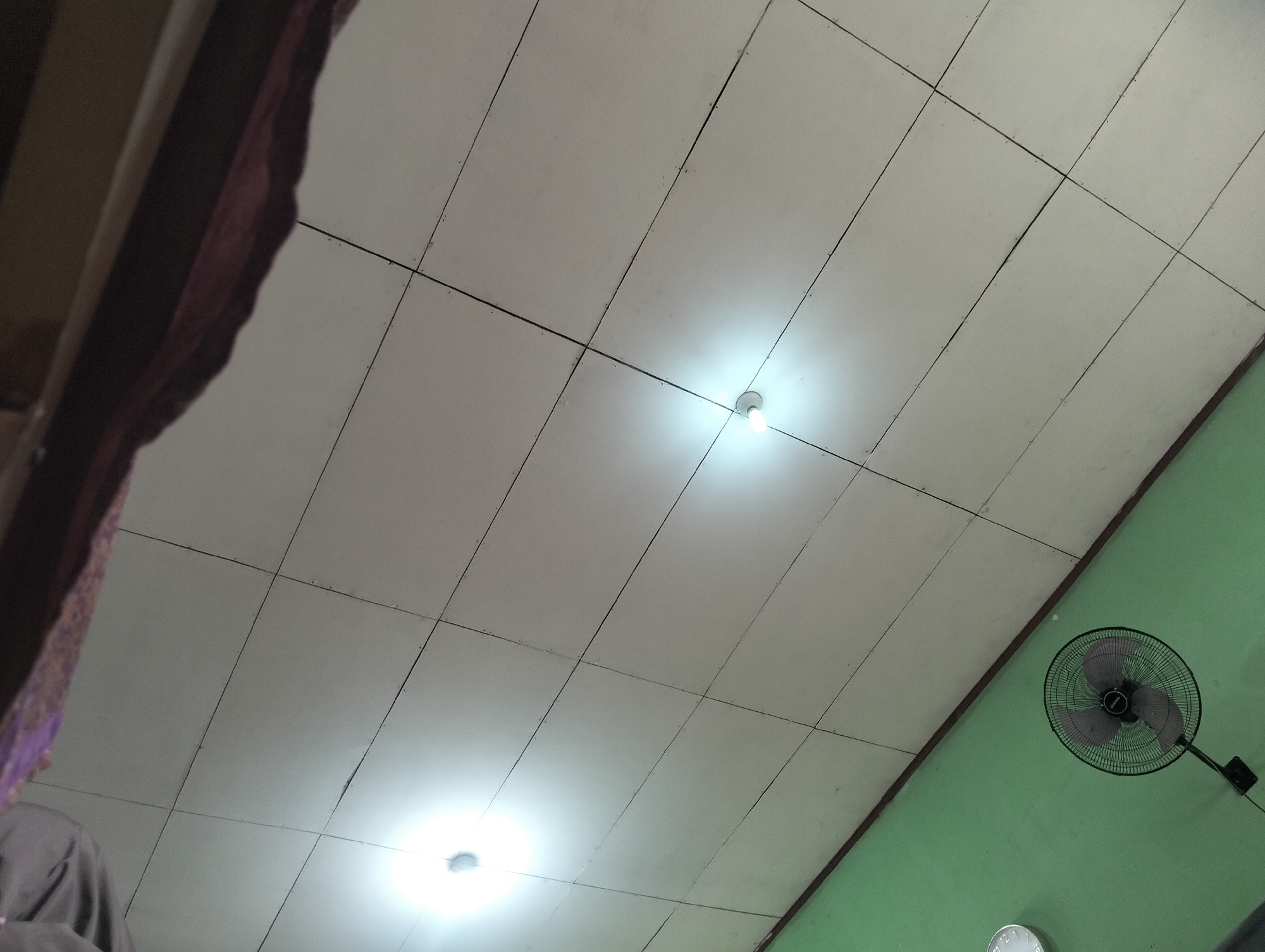 The image is a low-angle shot looking up at a tiled ceiling with two visible lightbulbs. To the right, a green wall is visible, along with a black oscillating fan mounted on it and a partial view of a clock. On the left side of the image, a dark, draped fabric is visible, obscuring the corner of the room. The scene appears to be indoors, possibly in a residential or commercial space in Banjar Agung, Indonesia. There are no people clearly depicted.