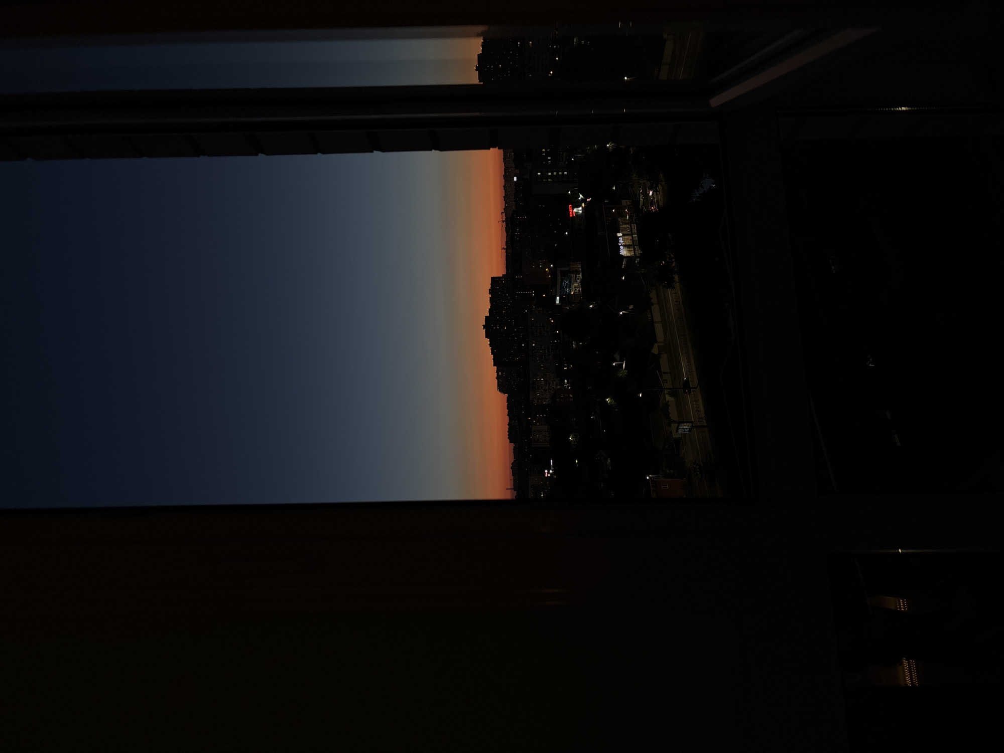 The image captures a twilight view of a city skyline, likely Krasnodar, Russia, from an elevated vantage point, possibly a window. The sky transitions from a deep navy blue at the top to a vibrant orange and red near the horizon, indicating the period shortly after sunset. Silhouetted against this colorful sky are numerous buildings of varying heights, with some appearing to be apartment complexes and others smaller structures. Streetlights and illuminated windows dot the cityscape, suggesting the transition from day to night. A distinct orange glow emanates from one building, possibly a commercial establishment. The foreground is dominated by dark, out-of-focus elements, which appear to be the interior of the room from which the photograph is taken, including a window frame. The overall atmosphere is one of urban tranquility as the city settles into the evening.